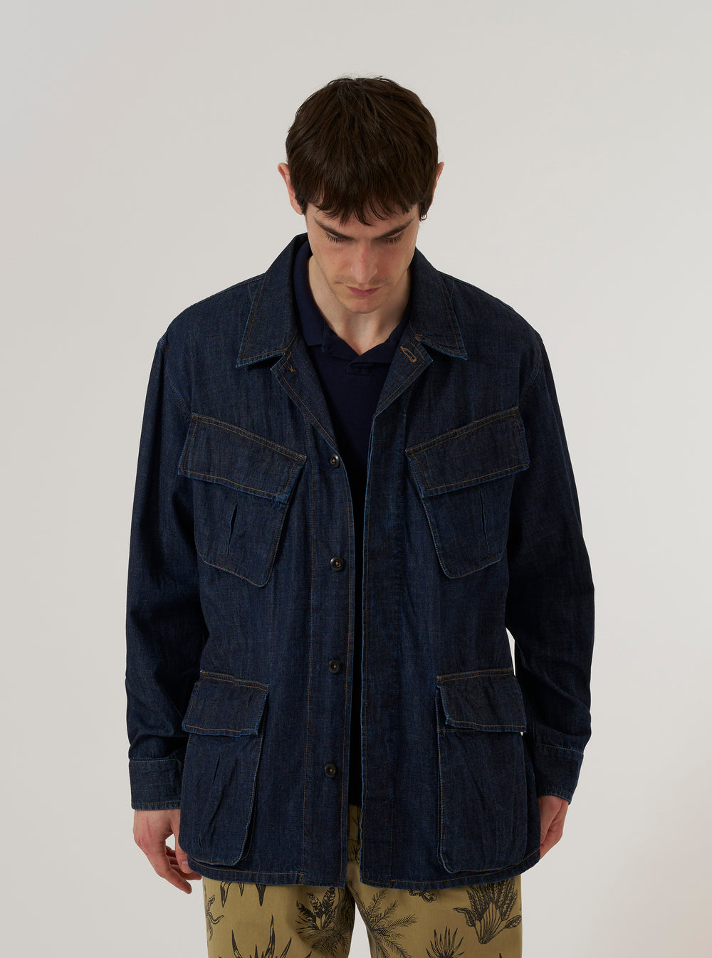 A model wears the Indigo Kaihara LT WT Denim Jungle Jacket over a light-colored top, standing against a plain white background. The jacket is crafted from deep indigo denim and features four large front patch pockets with button closures and a full-length buttoned front.