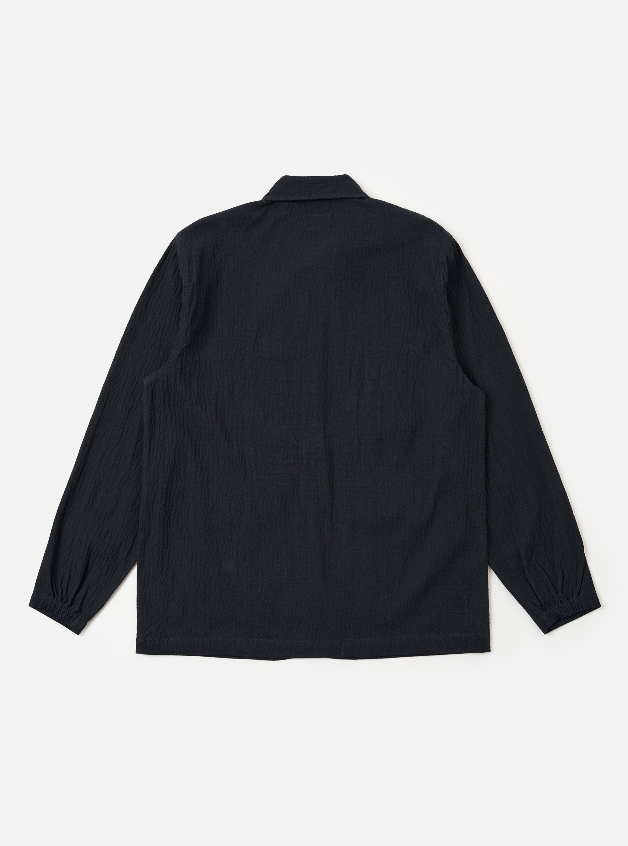The Dark Navy Ospina Cotton Labour Jacket is shown from the back as a flat lay against a plain white background. The image highlights the clean, simple construction of the garment's rear panel, the straight hem, and the half-elasticated cuffs. The dark navy fabric features a visible seersucker texture.