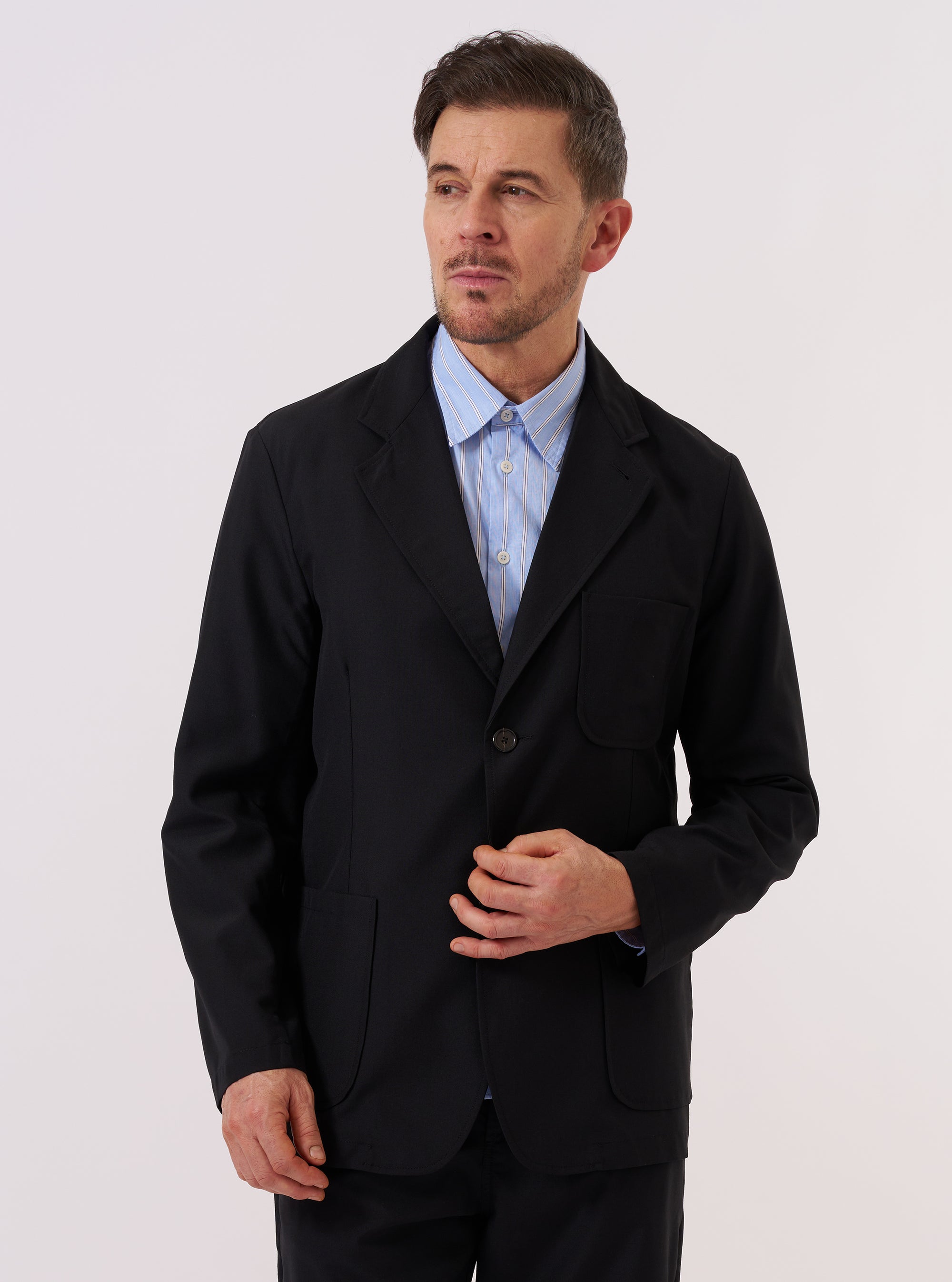 A model wears the Black Tropical Suiting Two Button Jacket over a light-colored shirt against a plain white background. The jacket features a classic two-button front closure, notched lapels, and a structured silhouette. The garment is made from a smooth, black fabric.