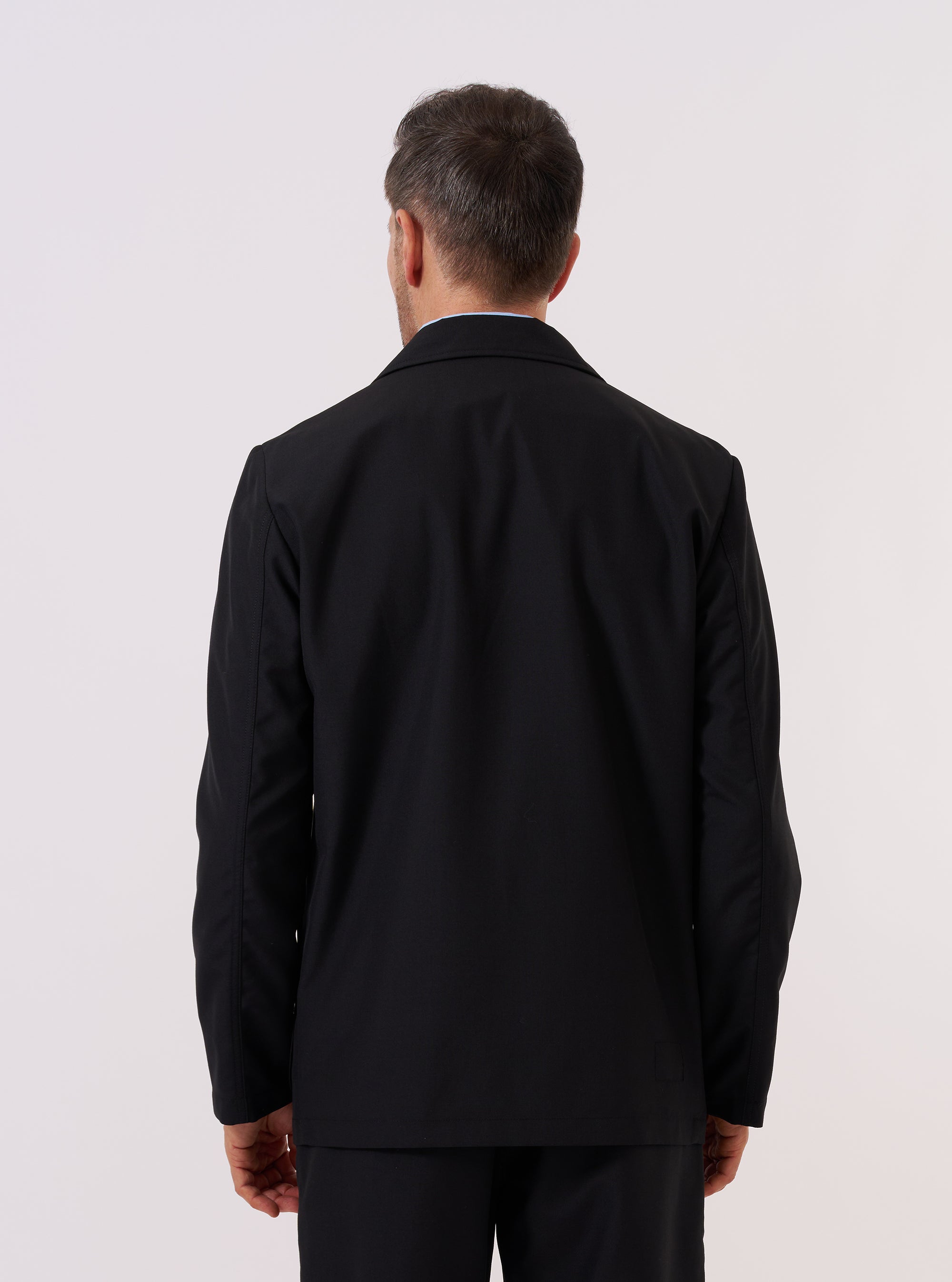 A model wears the Black Tropical Suiting Two Button Jacket over a dark top, shown from the back against a plain white background. The image emphasizes the jacket's unstructured silhouette, clean drape, and the two-piece sleeve construction. The smooth, lightweight texture of the tropical suiting fabric is visible. No mannequin is present in the image.
