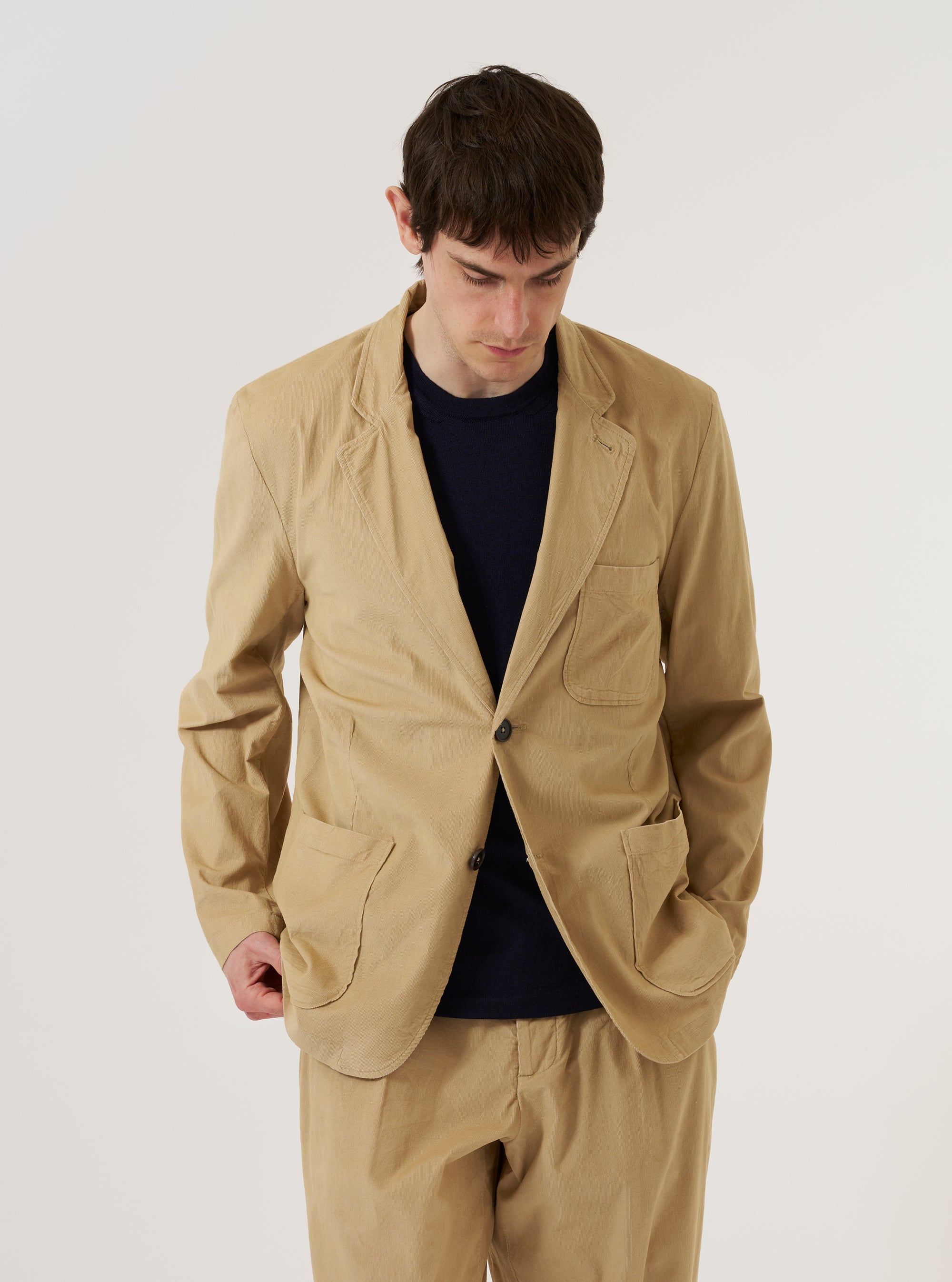 A model wears the Sand Organic Fine Cord Two Button Jacket over a light-colored shirt, standing against a plain, neutral background. The jacket features a classic two-button front closure, a notched lapel, and two large patch pockets at the waist. The sand-colored fabric has a visible fine corduroy texture.