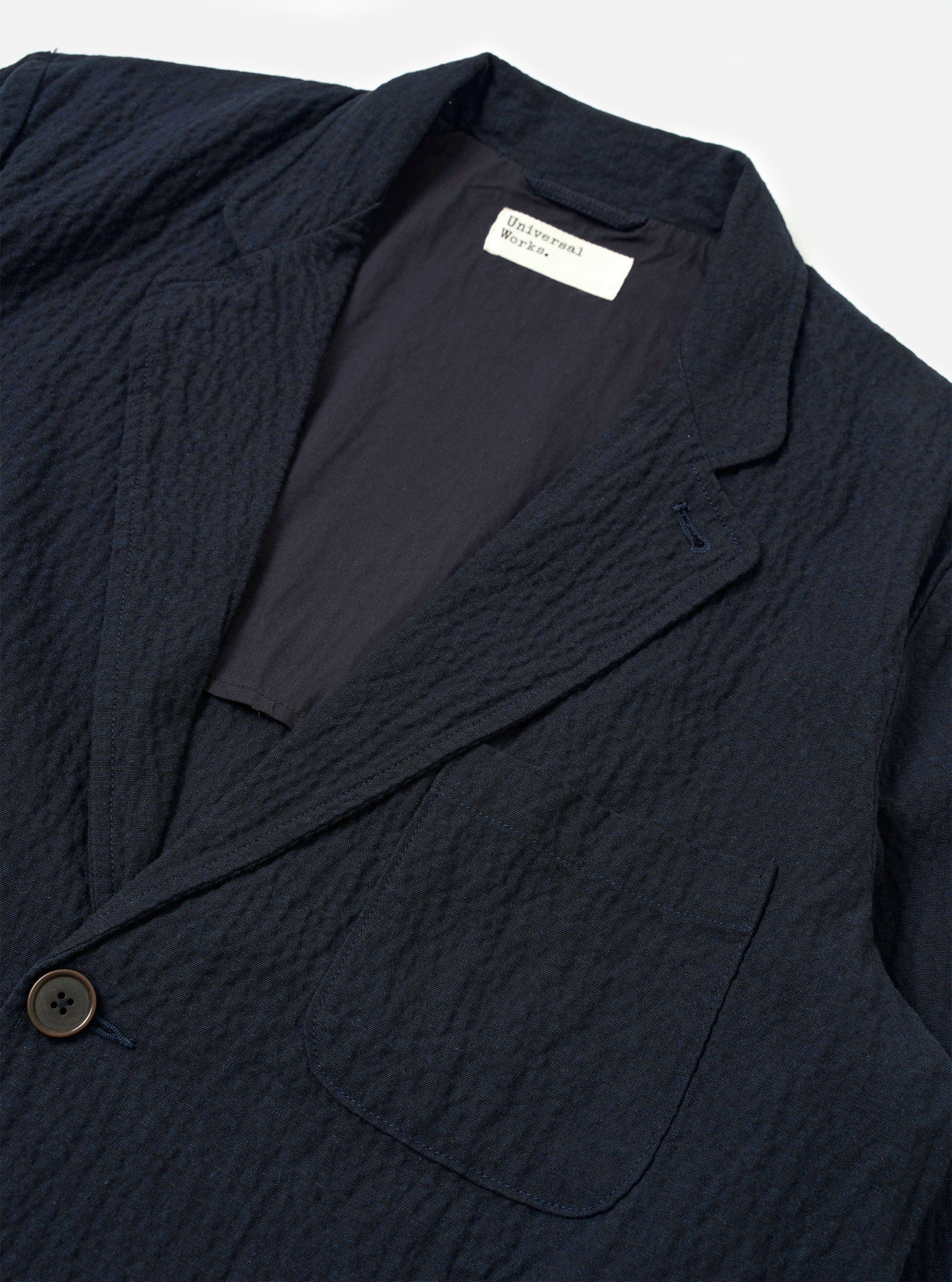 The Dark Navy Ospina Cotton Two Button Jacket is shown from a rear three-quarter angle as a flat lay against a plain white background. The image highlights the jacket's structured back with a central vertical seam, two-piece sleeve construction, and a straight hem. The dark navy fabric features a visible puckered seersucker texture.