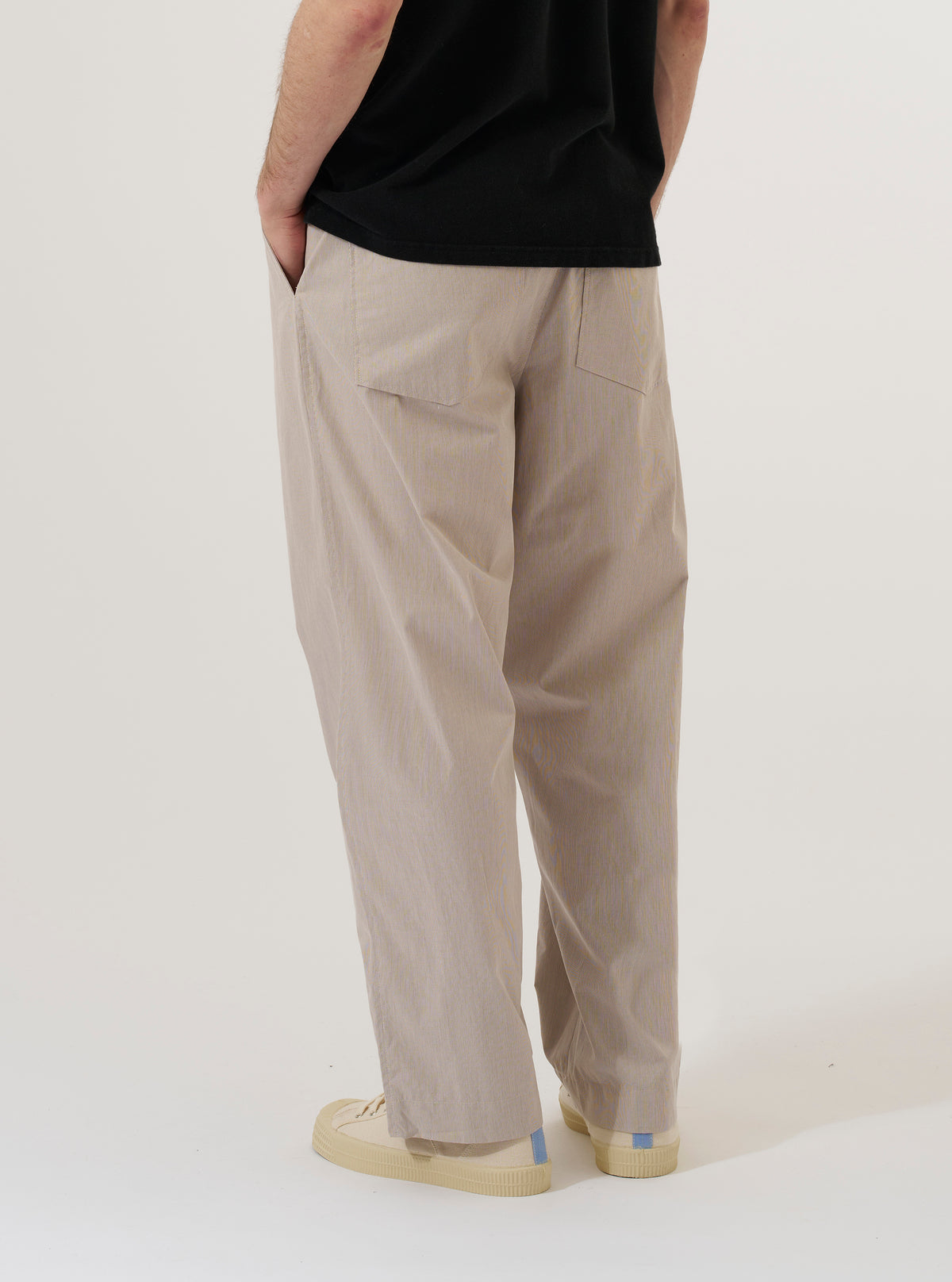 A model wears the Brown Kennedy Stripe Duke Pant, shown from the rear three-quarter angle against a plain white background. The trousers feature a loose, wide-leg fit with a brown and cream vertical stripe pattern. Visible details include two rear buttoned patch pockets and a straight hem. The model is wearing the pants with black shoes.