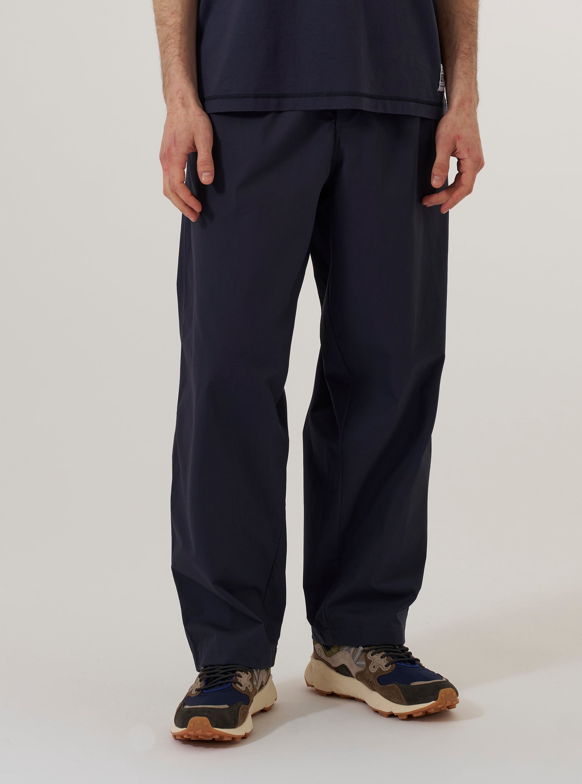 A model wears the Navy Peached Poplin Dutch Pant, shown from the waist down against a plain white background. The trousers feature a loose, wide-leg fit with a soft taper, double-pleated front, and slanted side pockets. The model is wearing the navy blue cotton trousers with a white T-shirt tucked into the waistband.