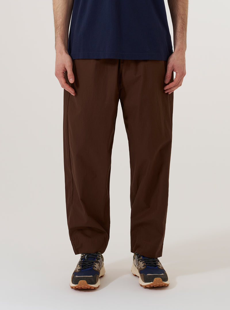 A model wears the Brown Peached Poplin Judo Pant against a plain white background. The image shows a front view of the trousers, highlighting the relaxed fit, elasticated drawstring waistband, and the smooth texture of the brown cotton poplin fabric. The model is wearing a light-colored top tucked into the pants, which feature a slightly tapered leg.