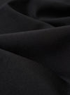 A close-up fabric swatch of the Black Tropical Suiting Military Chino highlights the smooth, technical texture and deep black color of the polyester and wool blend material. The fabric fills the entire frame, showcasing the fine weave of the tropical suiting.