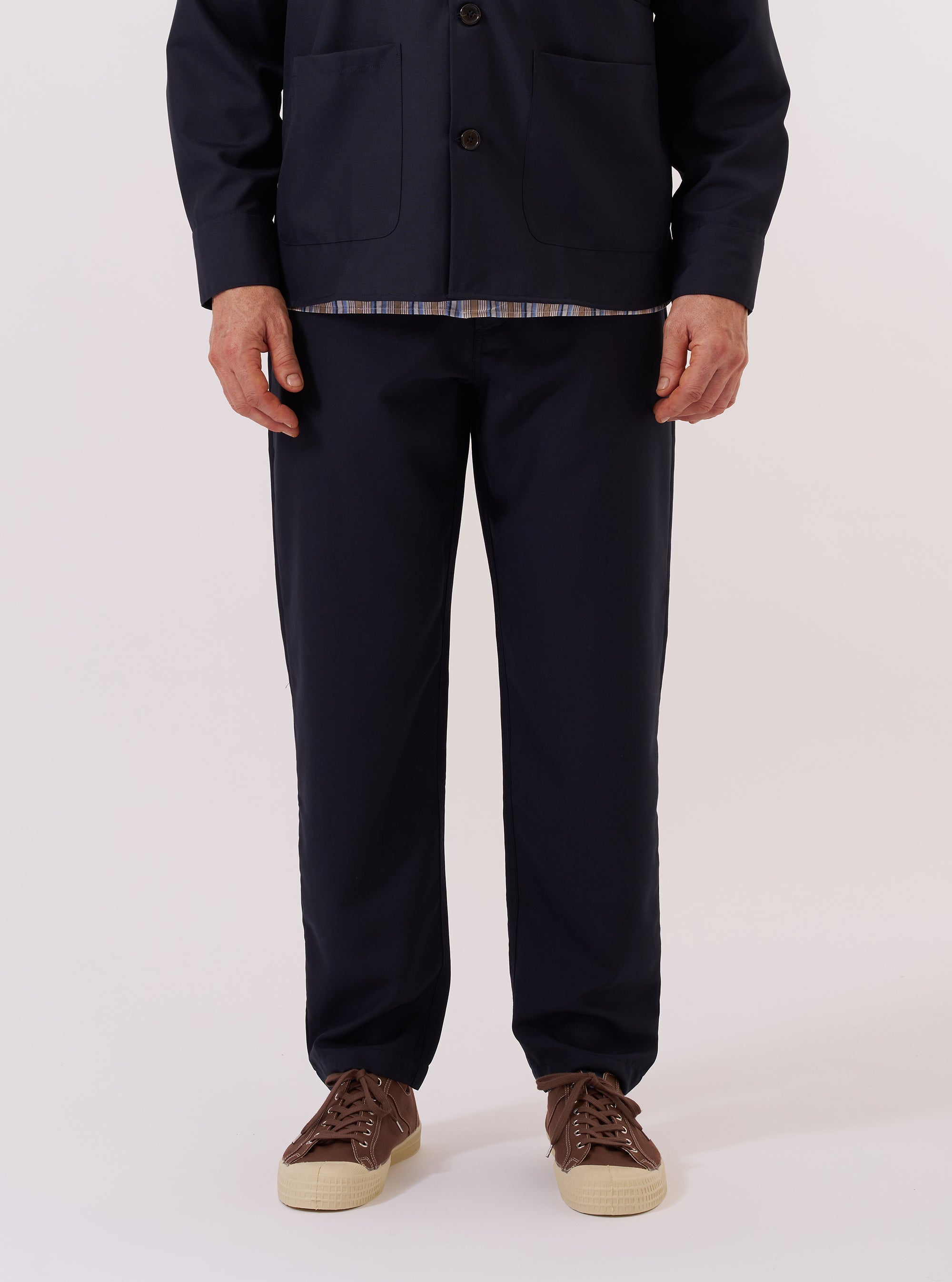 A model wears the Navy Tropical Suiting Military Chino against a plain white background. The image shows a front view of the trousers, highlighting the mid-rise waist, button fly, and two front pockets. The lightweight navy fabric has a clean, smooth appearance, and the leg shows a gradual taper towards the ankle. The model is wearing a light-colored shirt tucked into the chinos and white socks with loafers.