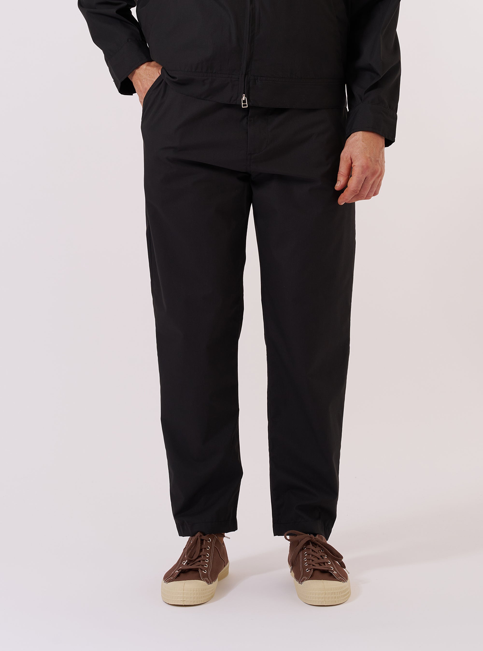 A model wears the Black Recycled Polytech Military Chino, shown from the waist down against a plain white background. The image highlights the slim-straight fit of the trousers, which feature a button fly, belt loops, and front pockets. The model is wearing the black chinos with a white t-shirt tucked in and white socks.