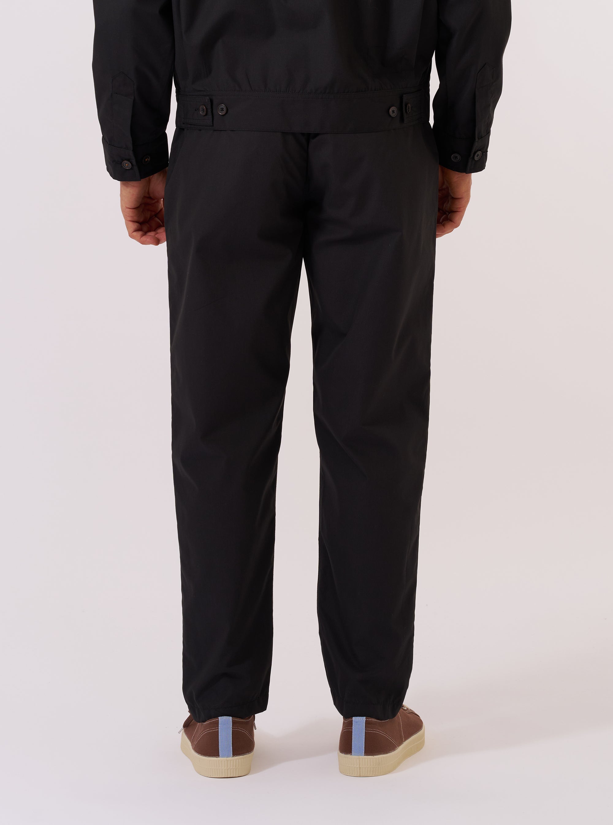 A model wears the Black Recycled Polytech Military Chino, shown from the waist down against a plain white background. The trousers feature a slim, straight-leg fit and are made from a smooth black recycled polytech fabric. The image highlights the clean, military-style construction and the technical texture of the material.