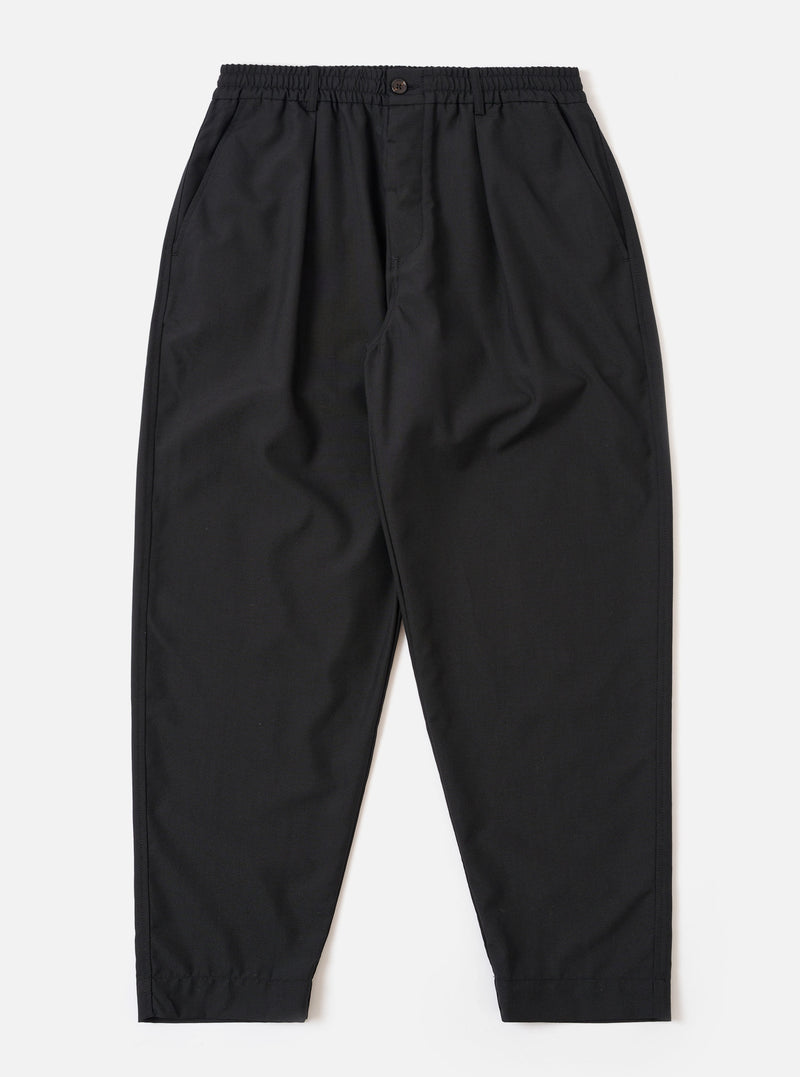 The Black Tropical Suiting Pleated Track Pant is shown as a flat lay against a plain white background. The trousers feature an elasticated waistband with an internal drawstring, a single front pleat on each leg, and two side pockets. The black tropical suiting fabric is a lightweight polyester and wool blend with a smooth finish.