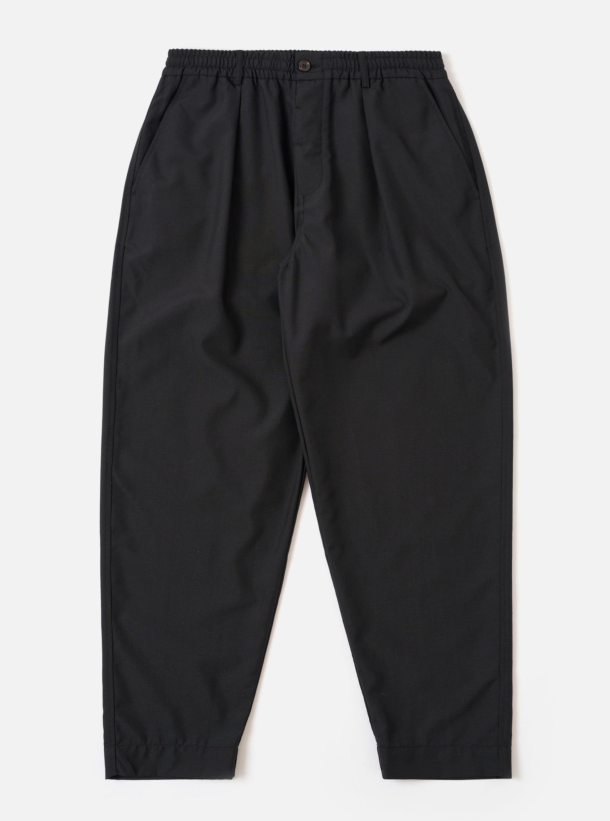 The Black Tropical Suiting Pleated Track Pant is shown as a flat lay against a plain white background. The trousers feature an elasticated waistband with an internal drawstring, a single front pleat on each leg, and two side pockets. The black tropical suiting fabric is a lightweight polyester and wool blend with a smooth finish.