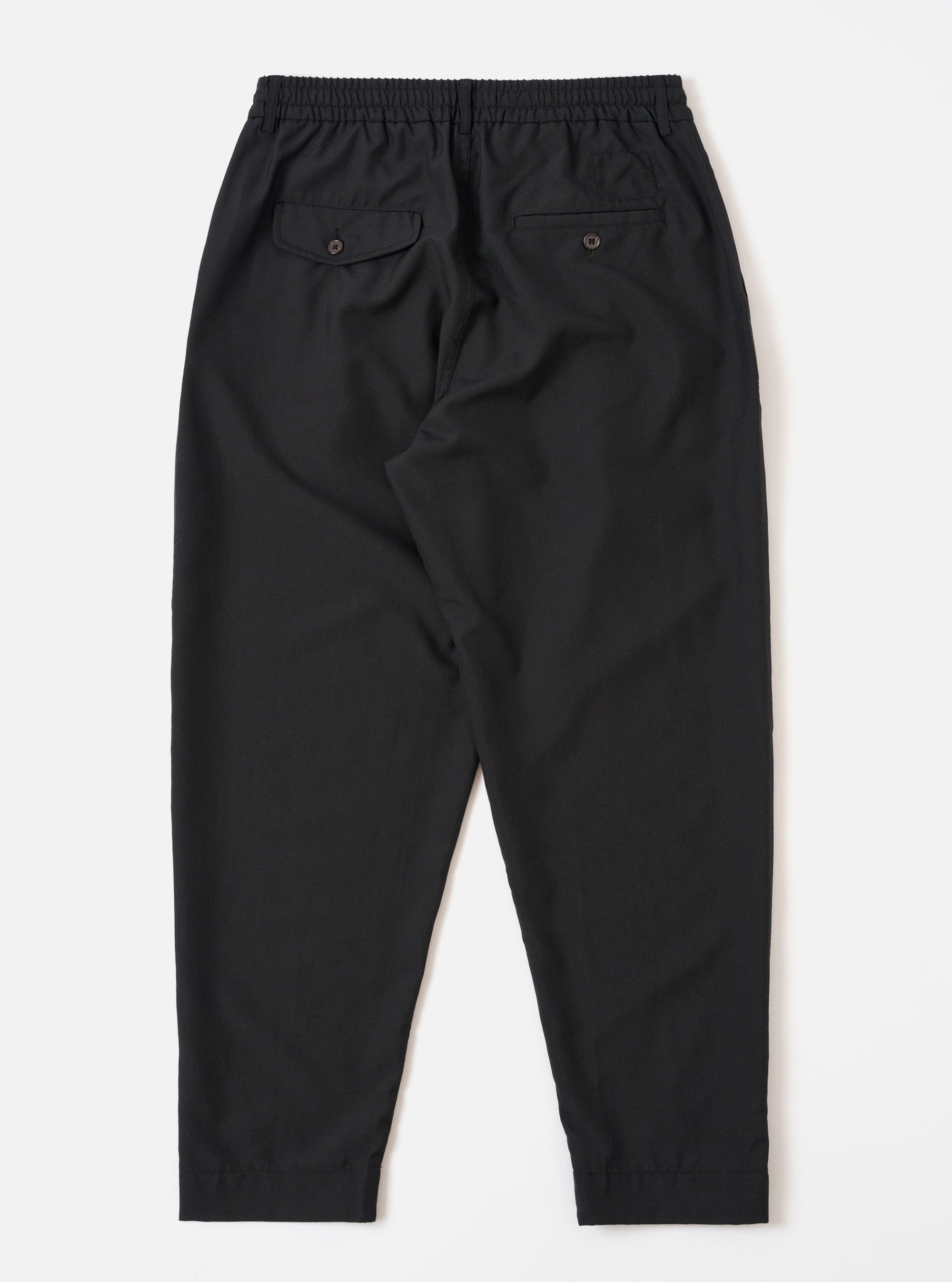 The Black Tropical Suiting Pleated Track Pant is shown from the back as a flat lay against a plain white background. The image highlights the clean construction of the trousers, featuring two rear pockets: one buttoned flap pocket on the left and one button jetted pocket on the right. The black tropical suiting fabric appears smooth and lightweight.