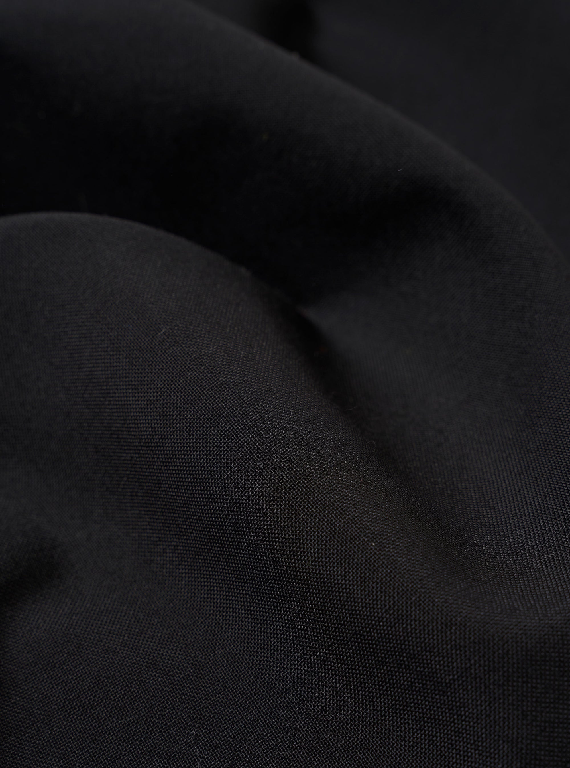 A close-up fabric swatch of the Black Tropical Suiting Pleated Track Pant is shown, highlighting the fine, smooth texture and solid black color of the lightweight tropical suiting material.
