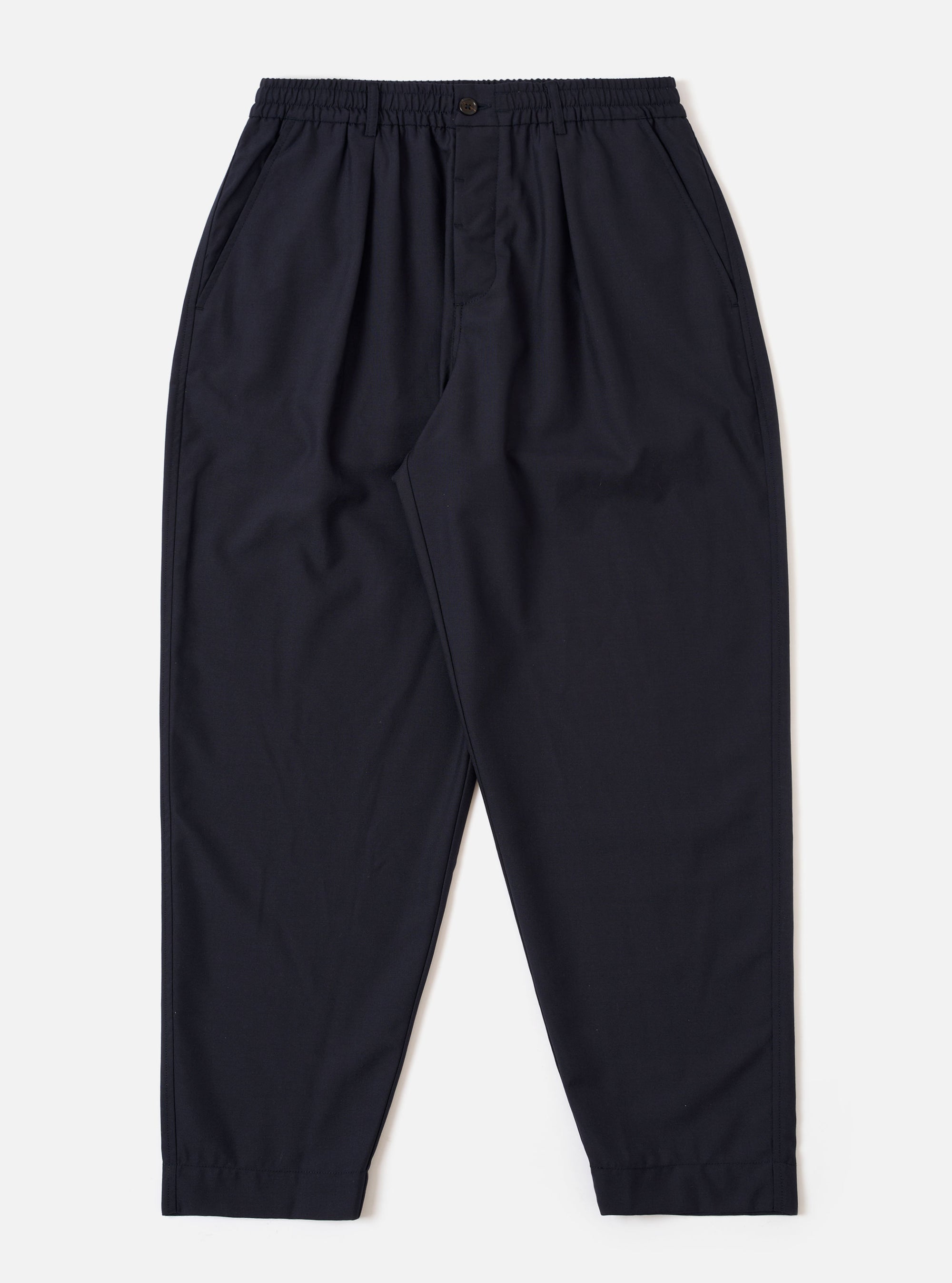 The Navy Tropical Suiting Pleated Track Pant is shown as a flat lay against a plain white background. The trousers feature an elasticated waistband with a tonal drawstring, a single pleat at the front of each leg, and a tapered silhouette. The fabric is a smooth, lightweight navy blue tropical suiting.
