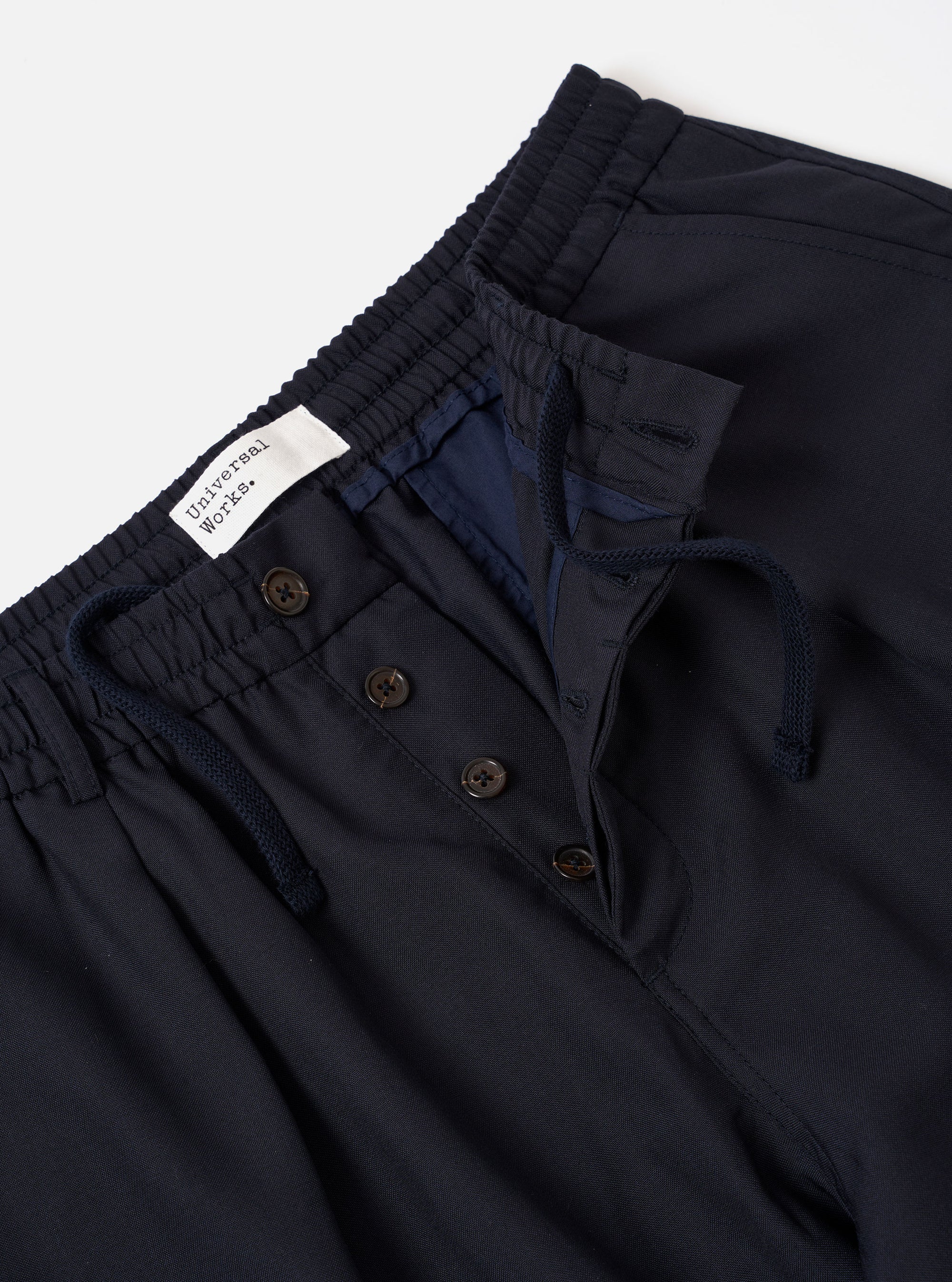 The Navy Tropical Suiting Pleated Track Pant is shown from the back as a flat lay against a plain white background. The image highlights the two rear set-in pockets, one with a buttoned flap and one button-jetted, as well as the elasticated waistband. The fabric is a smooth, dark navy tropical suiting with a visible fine-weave texture.