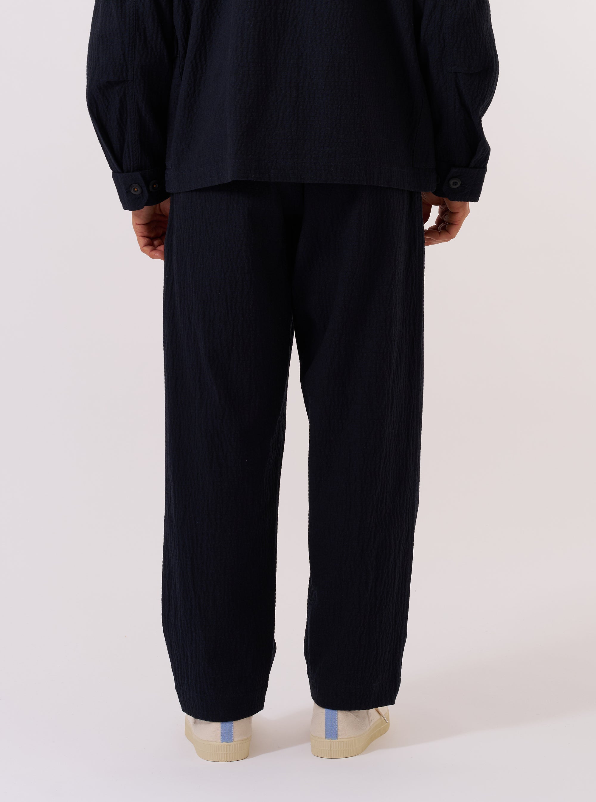 A model wears the Dark Navy Ospina Cotton Oxford Pant, shown from a rear three-quarter angle against a plain white background. The image highlights the loose, wide-leg fit of the trousers and the puckered seersucker texture of the dark navy fabric. The model is wearing the pants with a matching dark navy jacket and white shoes.