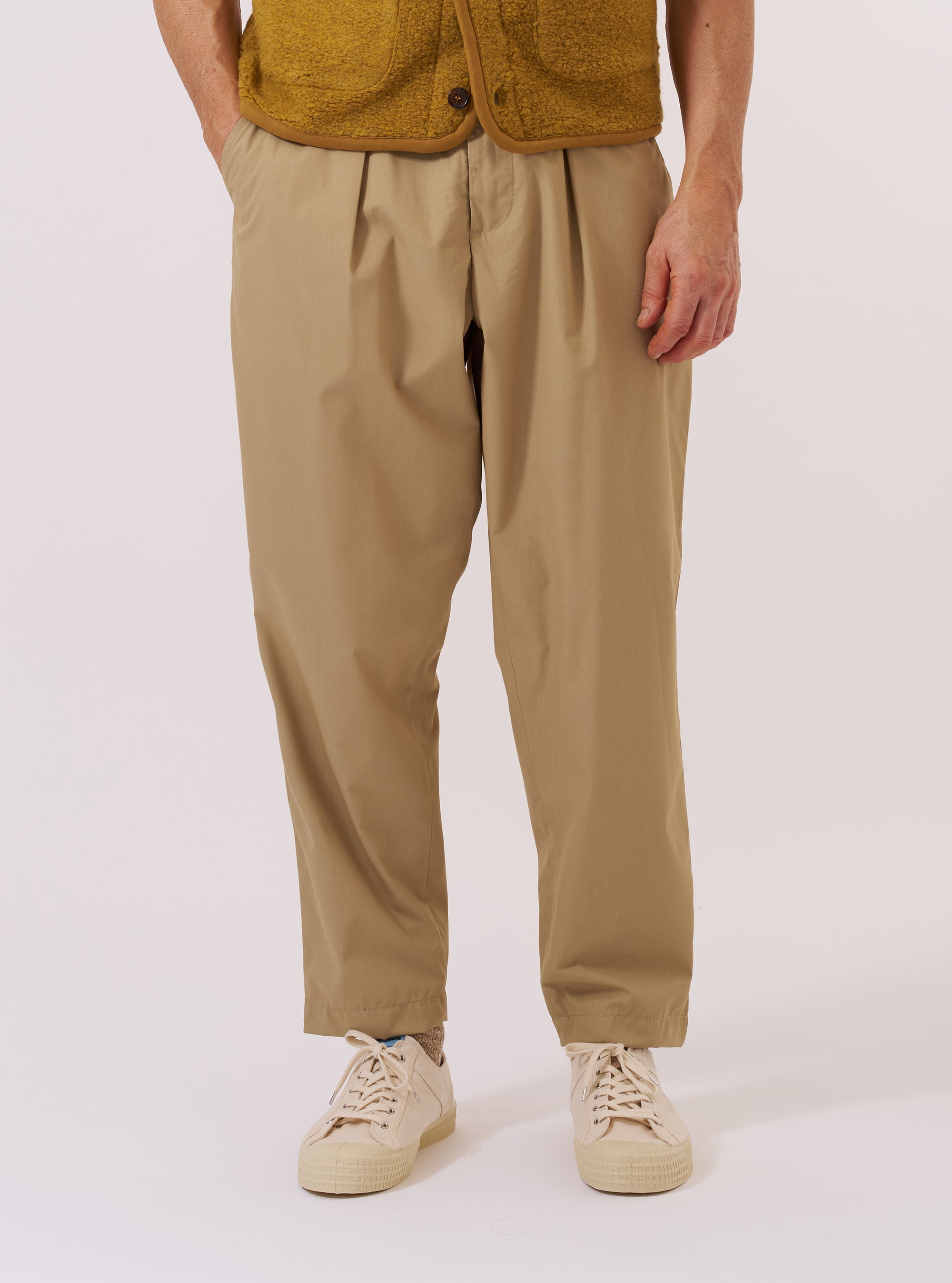 A model wears the Sand Recycled Polytech Pleated Track Pant while standing against a plain white background. The image shows a front view of the trousers, highlighting the loose fit, tapered leg, and the single pleat at the front. The elasticated waistband and the light sand-colored recycled poly-tech fabric are prominent. The model is wearing the trousers with a light-colored top tucked in to display the waistband detail.