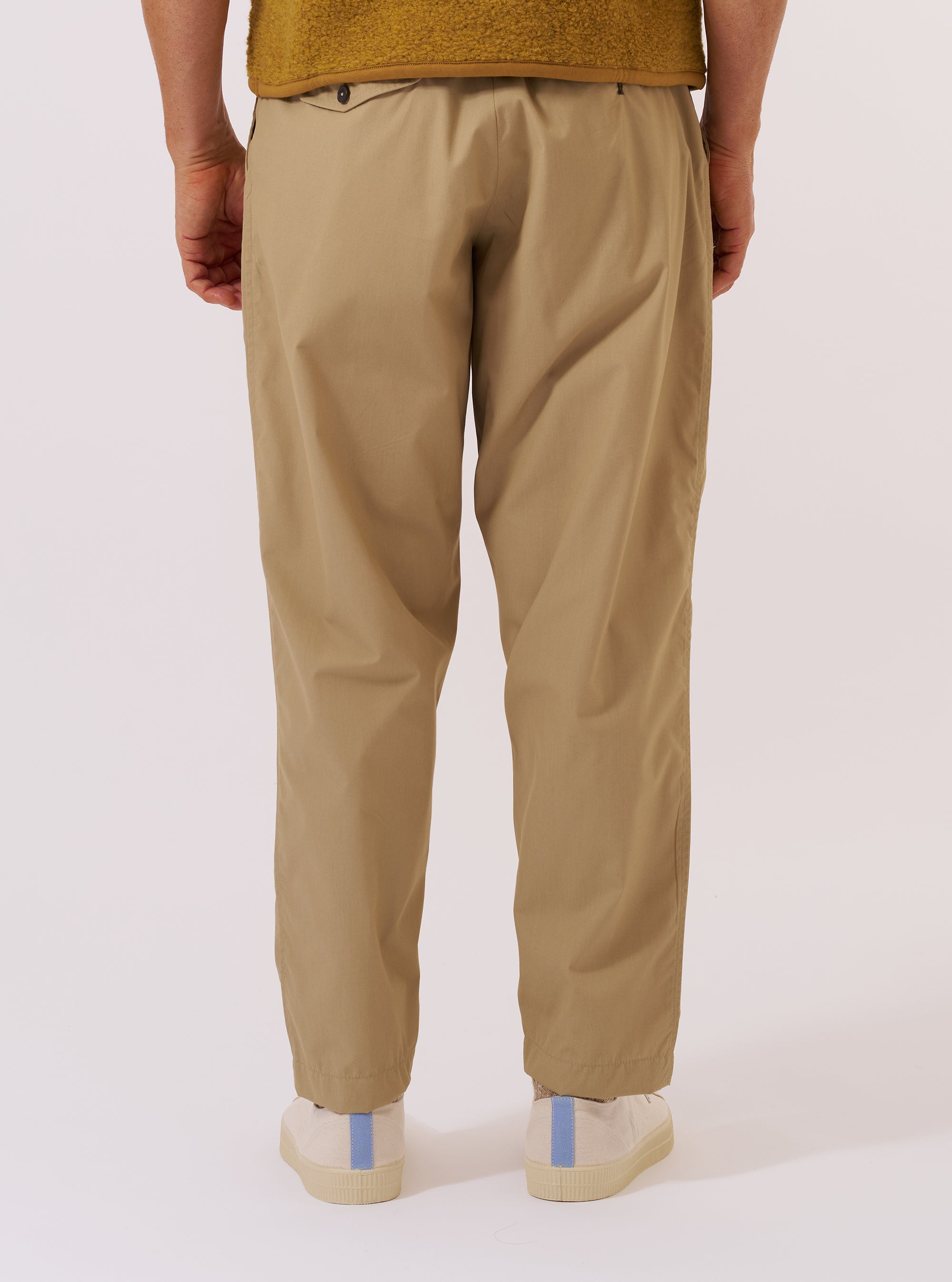 A model wears the Sand Recycled Polytech Pleated Track Pant, shown from the waist down against a plain white background. The image highlights the loose, tapered fit of the trousers, the single pleat at the front, and the elasticated waistband with a visible drawstring. The model is also wearing white socks and dark-colored shoes.
