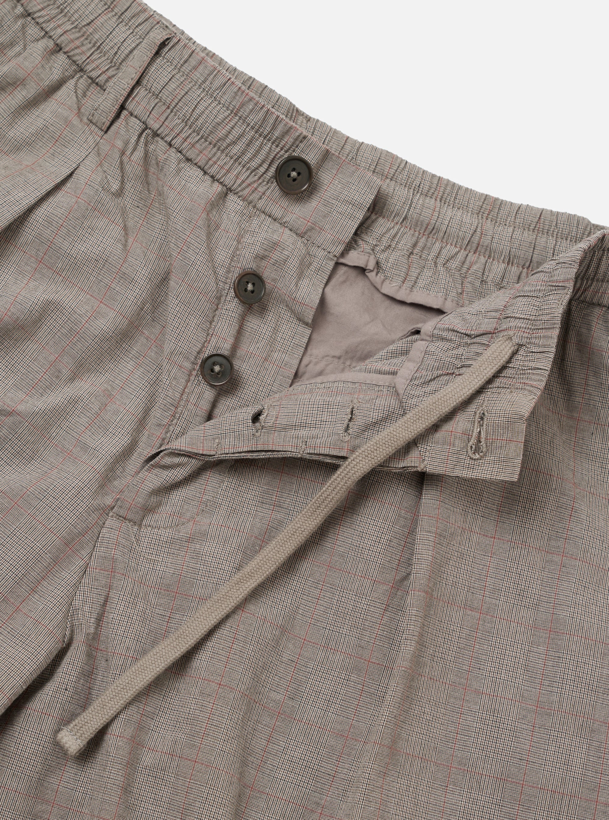 The Grey Brown Wales Glen Check Pleated Track Pant is shown as a flat lay against a white background. The trousers feature a grey and brown Glen check pattern, an elasticated waistband with a drawstring, and a single front pleat on each leg. The image highlights the loose, tapered fit and the texture of the cotton fabric.