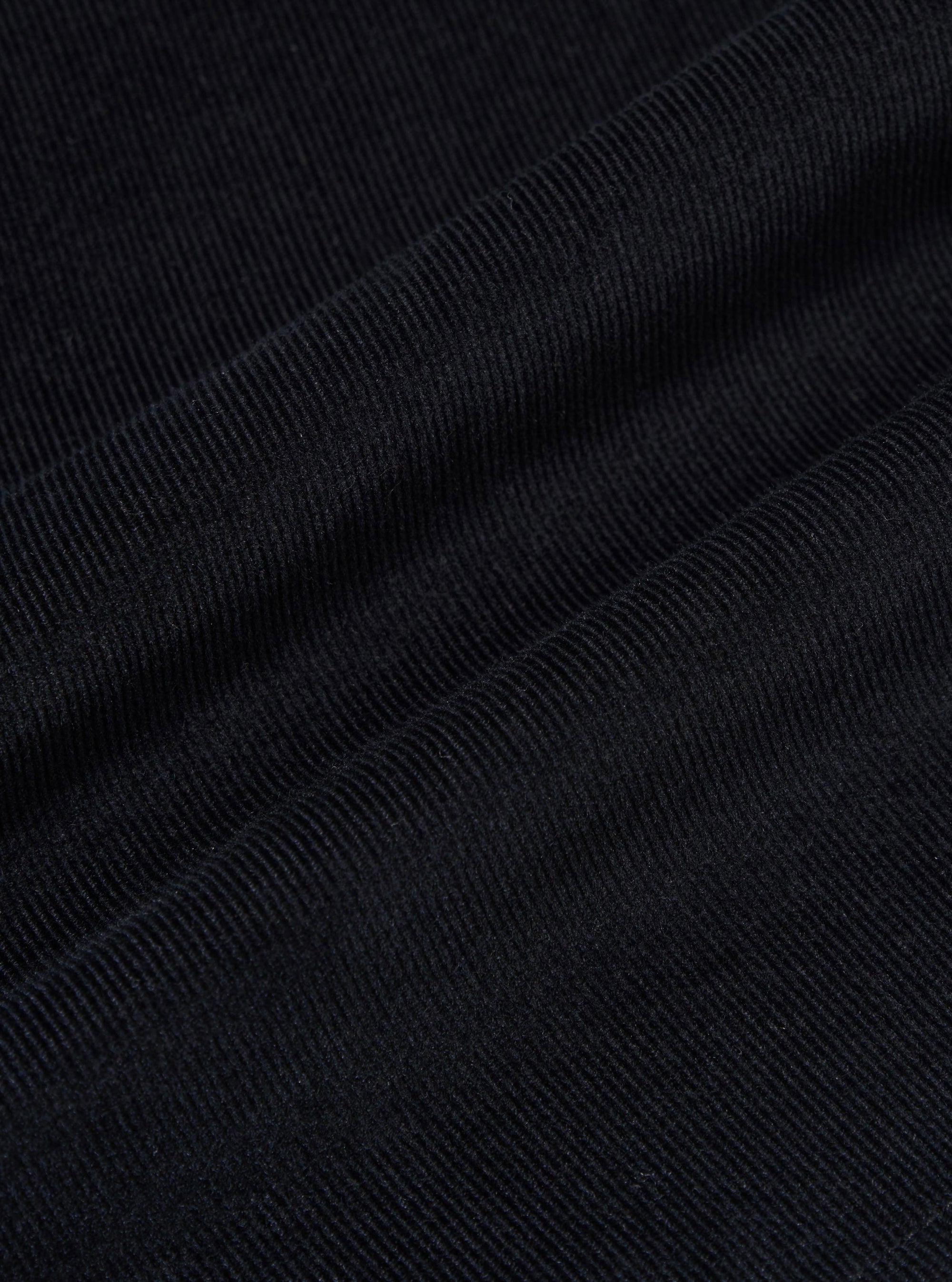A close-up fabric swatch of the Black Organic Fine Cord Fatigue Short highlights the fine-ribbed texture and soft finish of the organic cotton corduroy. The image focuses on the deep black color and the vertical lines of the thin wales.