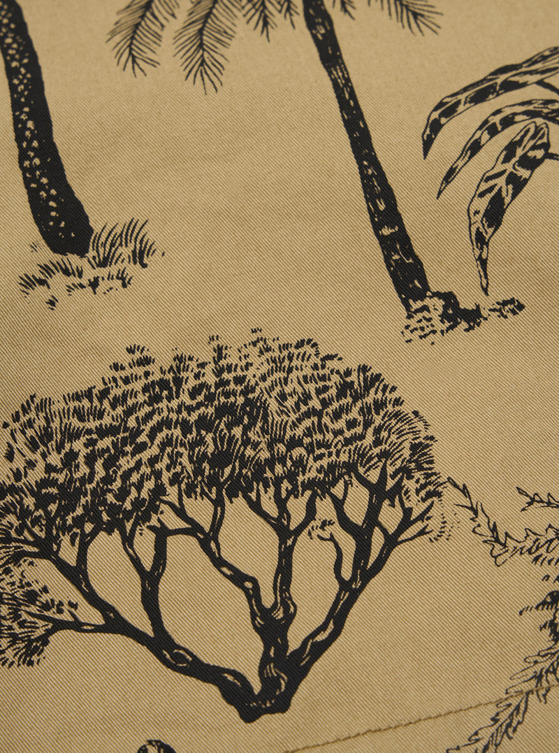 A close-up swatch of the Sand Jungle Toile Twill Pleated Track Short highlights the texture and intricate pattern of the fabric. The design features a dark jungle-themed toile print including trees and foliage against a light sand-colored base. The image provides a detailed view of the cotton twill weave.