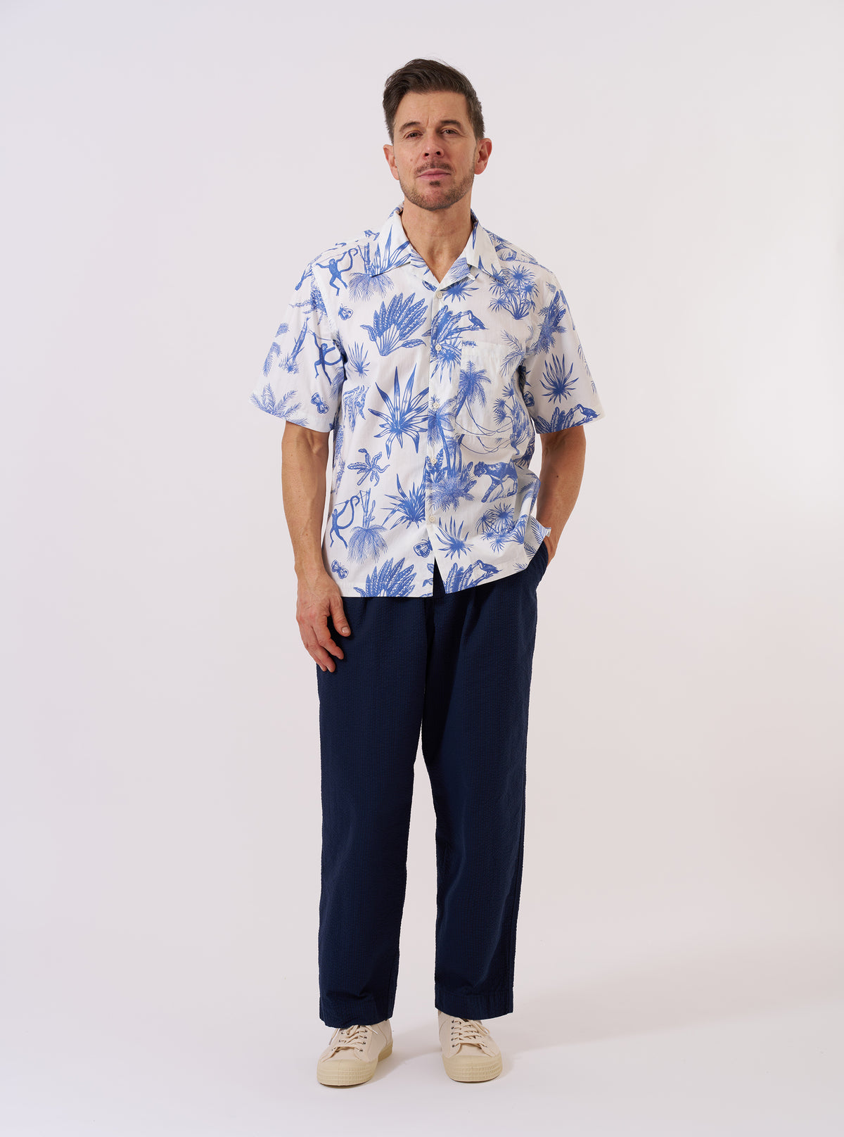 The White Jungle Toile Poplin Camp Shirt is shown as a flat lay against a white background. The shirt features a camp collar, a single chest patch pocket, and a central button-down front. The fabric is a white cotton poplin decorated with a detailed jungle toile print featuring animals and tropical foliage in a dark ink.