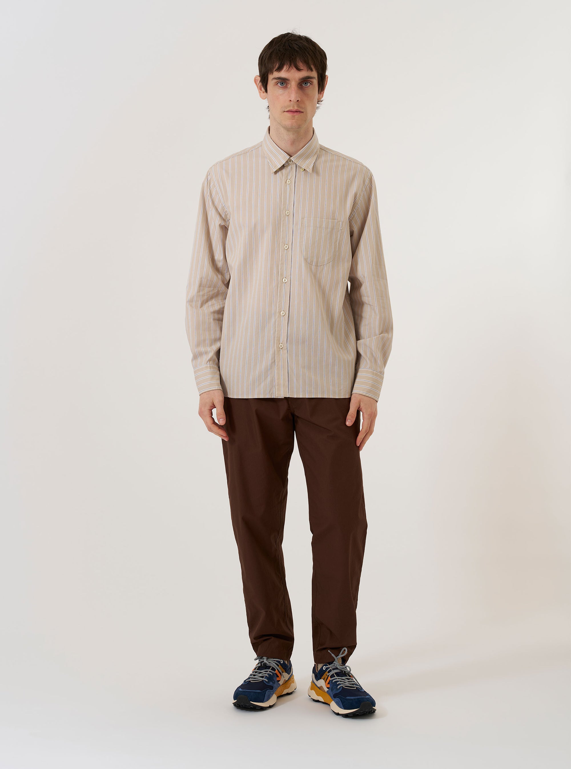 The Sand Urban Stripe Lazy Day Shirt is shown as a flat lay against a white background. The shirt features a button-down collar, a central button-down front, and a single asymmetric chest pocket. The fabric is a light sand color with thin, vertical urban stripes throughout.
