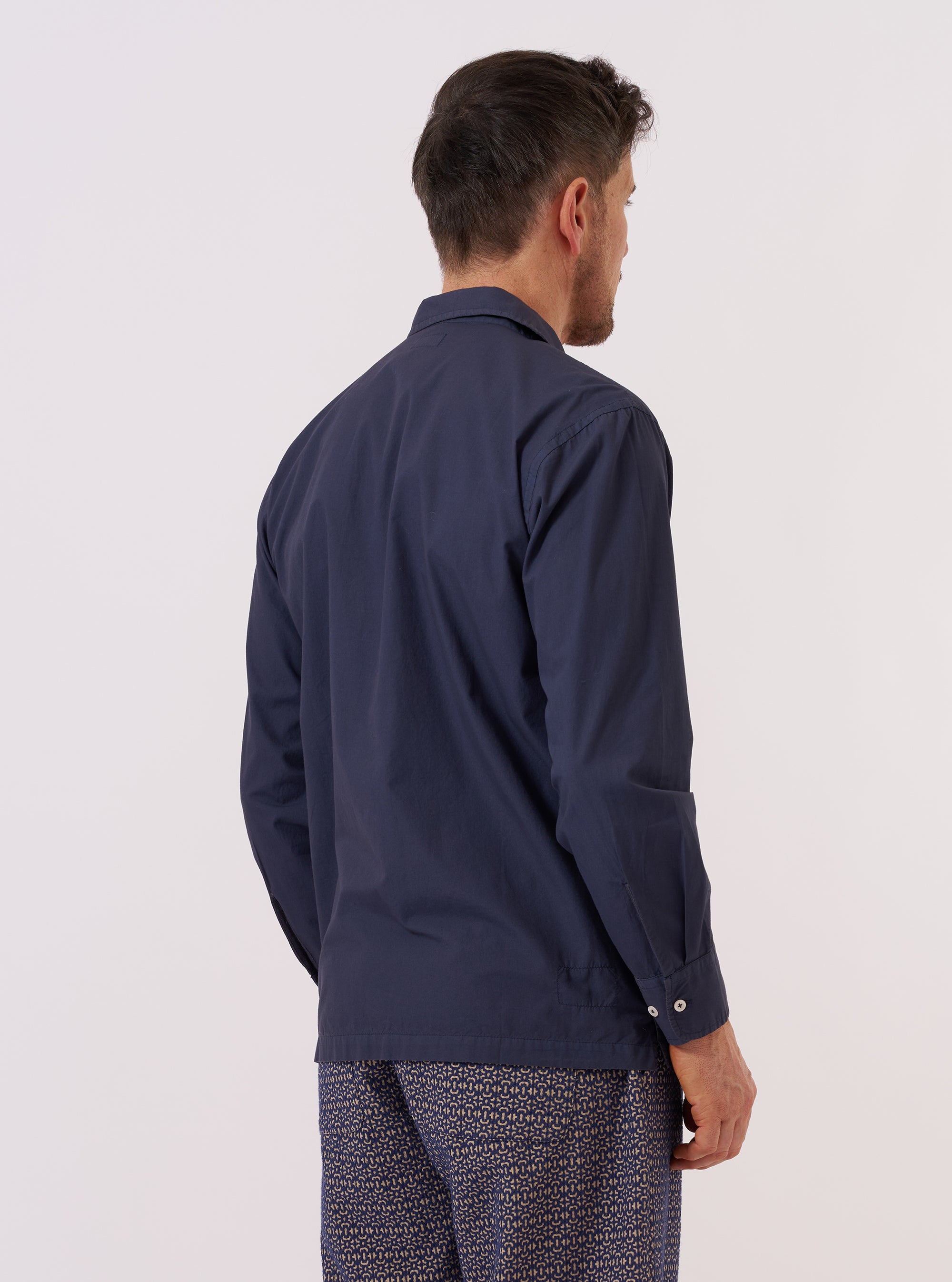 A model wears the Navy Poplin Dwight Overshirt while standing against a plain white background. The overshirt is featured in a front-facing view and includes a camp collar and two large utility-style chest pockets. It is worn over a light-colored t-shirt, highlighting the smooth texture of the navy blue cotton poplin fabric.
