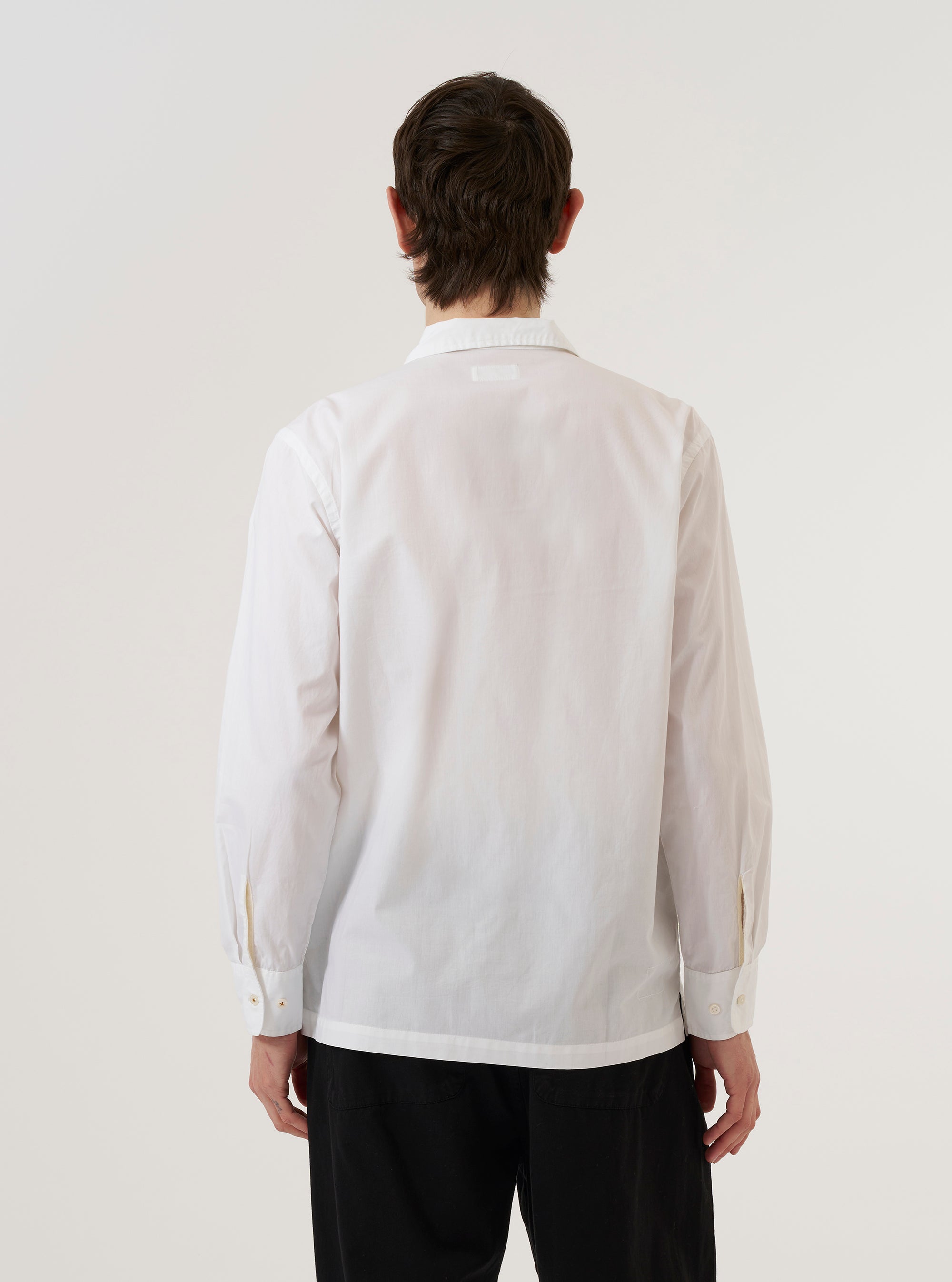 A model wears the White Poplin Dwight Overshirt against a plain white background, shown from a rear three-quarter angle. The image highlights the garment's clean, white poplin fabric, a straight hem, and a simple, structured back. The overshirt features a relaxed fit and a camp collar. No mannequin is present in the image.