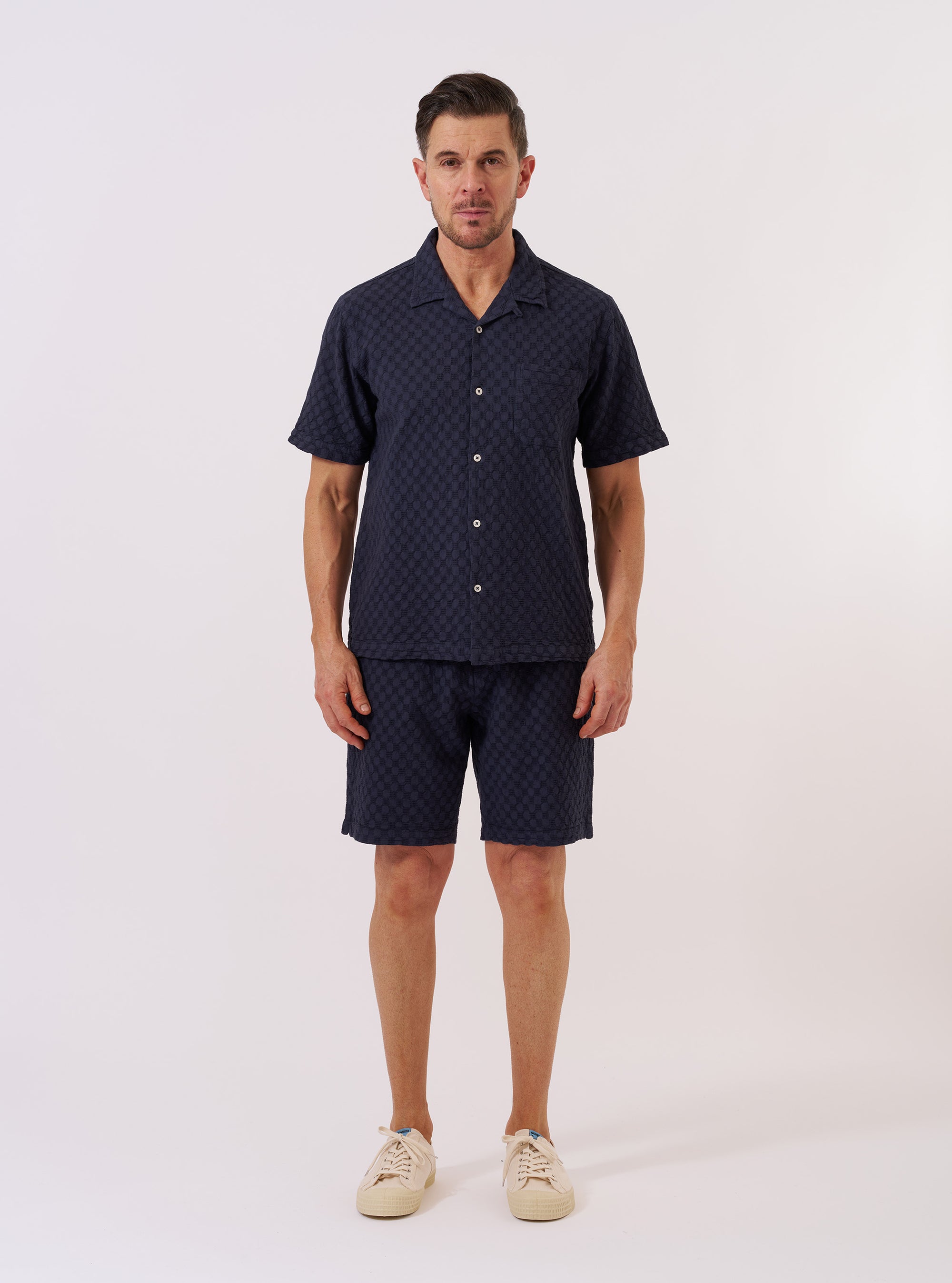 The Navy Dot Waffle Road Shirt is shown as a flat lay against a plain white background. The shirt features a camp collar, a single chest patch pocket, and a central button-down front with mother-of-pearl buttons. The navy blue cotton fabric has a distinct rounded waffle-dot texture throughout.