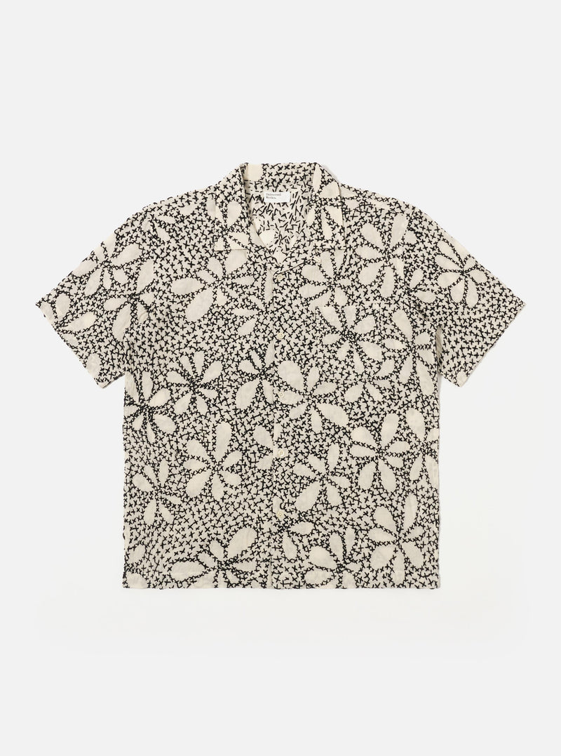 The Ecru/Black Kantha Flower Road Shirt is shown as a flat lay against a white background. The short-sleeve shirt features a camp collar, a central button-down front, and a single patch pocket on the left chest. The ecru fabric is detailed with an all-over black floral embroidery pattern.