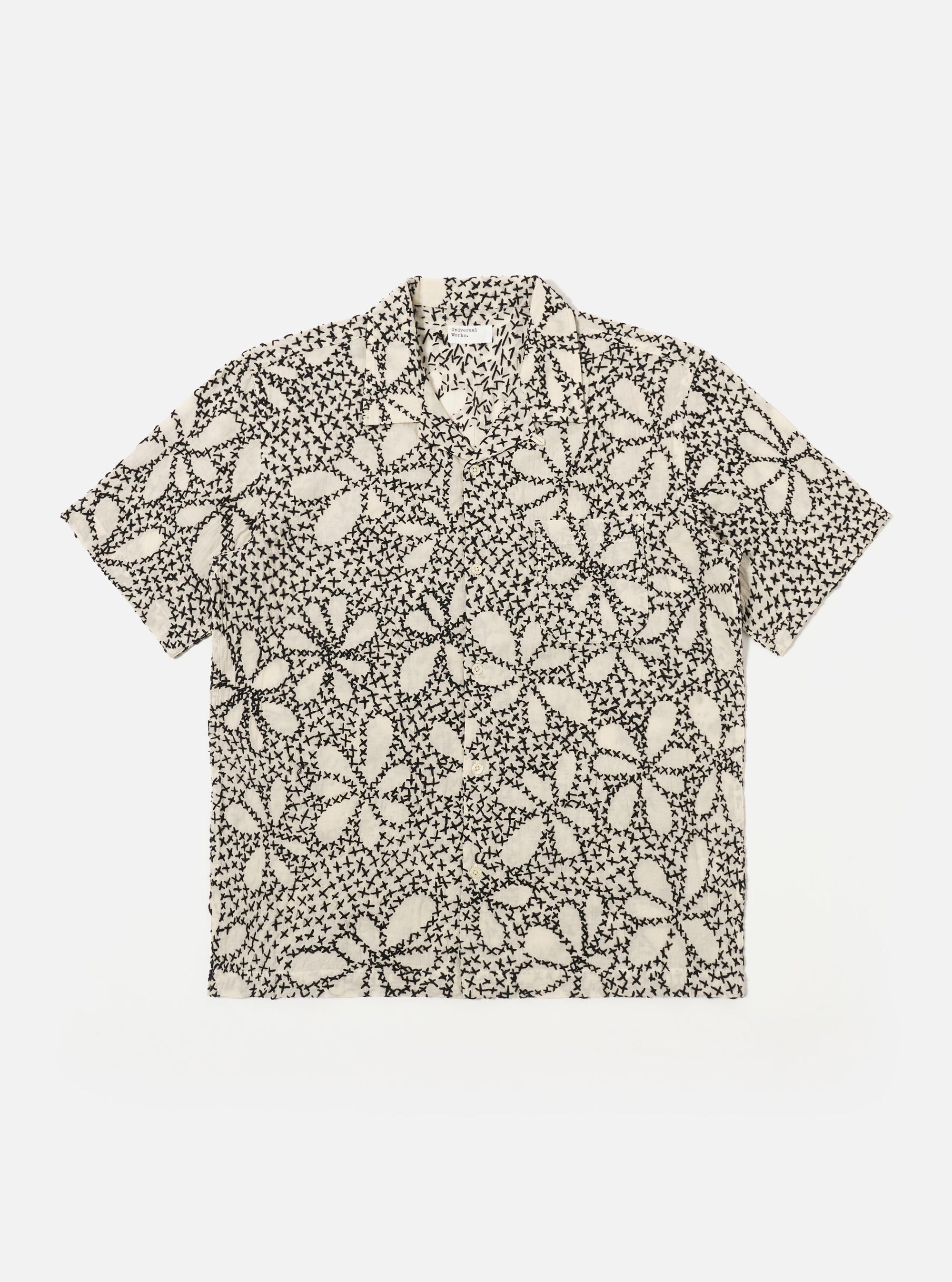 The Ecru/Black Kantha Flower Road Shirt is shown as a flat lay against a white background. The short-sleeve shirt features a camp collar, a central button-down front, and a single patch pocket on the left chest. The ecru fabric is detailed with an all-over black floral embroidery pattern.