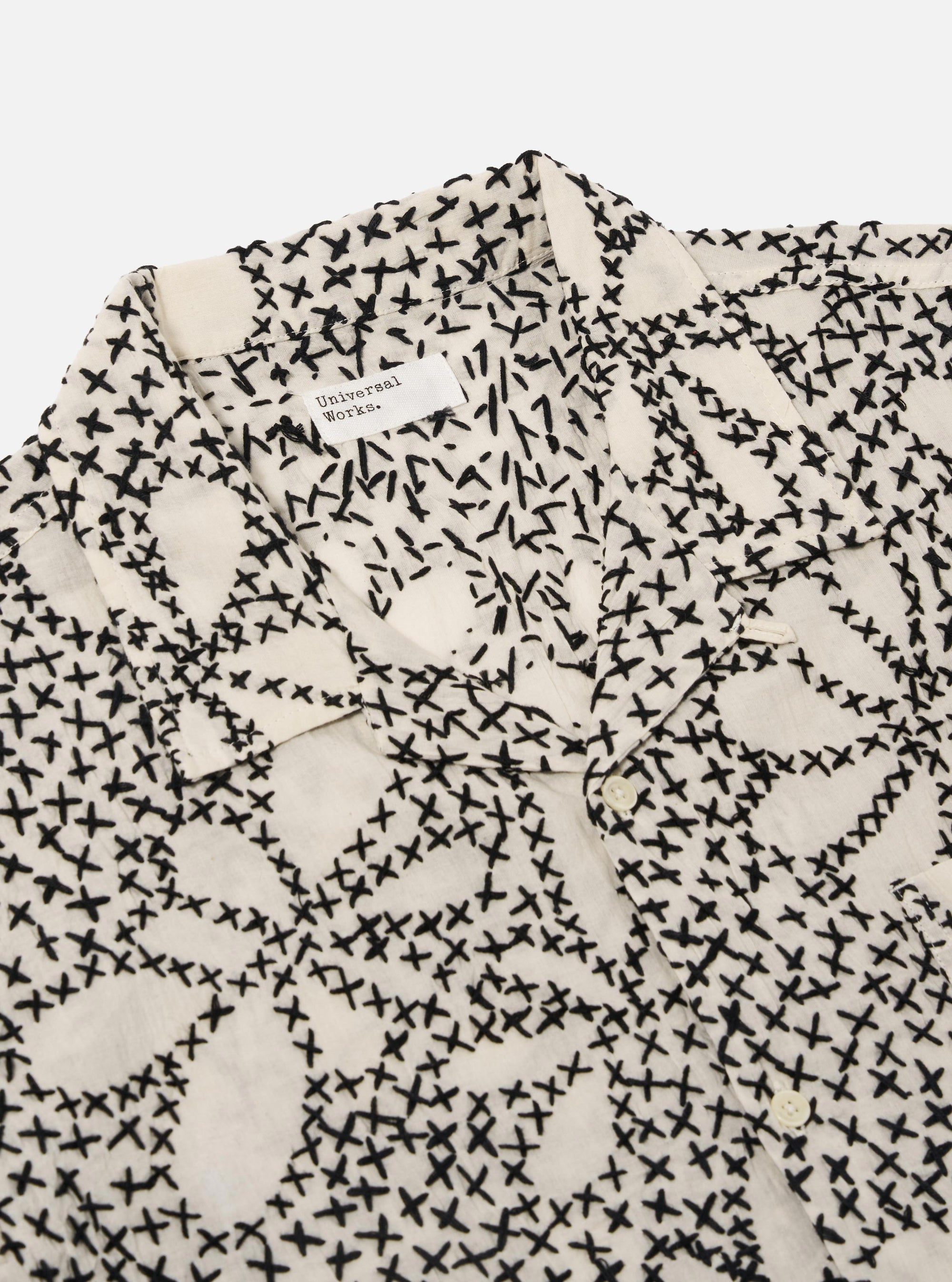 The Ecru/Black Kantha Flower Road Shirt is shown from a rear angle as a flat lay against a plain white background. The shirt features a straight hem and short sleeves, showcasing the intricate black floral Kantha embroidery across the ecru cotton fabric.