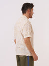 A model wears the Ecru 3D Flower Road Shirt against a neutral studio background. The shirt features a one-piece camp collar, a button-through front, and a single chest patch pocket. The garment is constructed from cotton fabric with a prominent 3D tufted flower texture throughout. The model is shown from the waist up, wearing the shirt with the top button open.