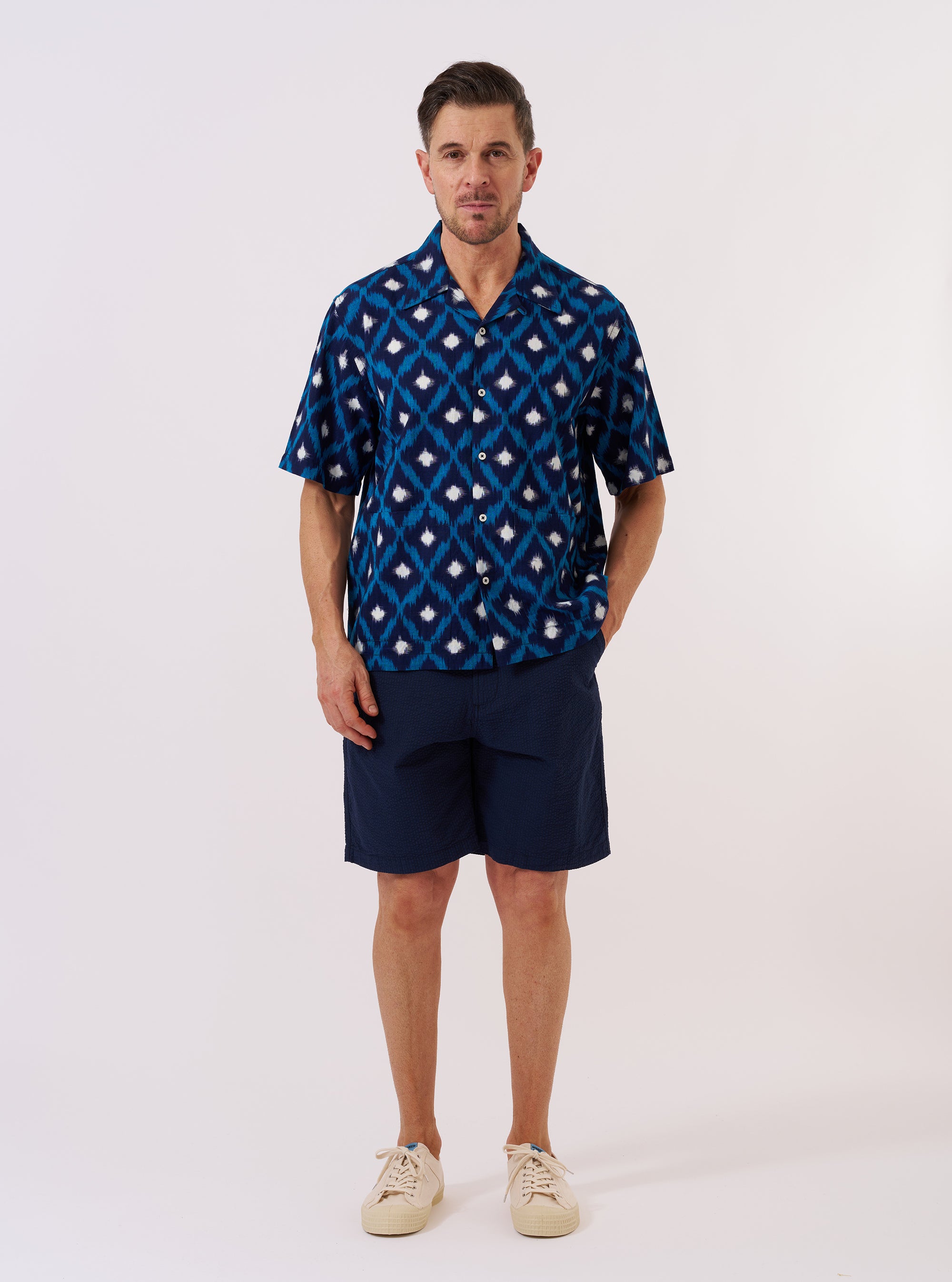 The Indigo Diamond Ikat Deck Shirt is shown as a flat lay against a plain white background. The shirt features short sleeves, a one-piece collar, a five-button front closure, and two large patch pockets at the lower front. The fabric displays an intricate repeating diamond ikat pattern in indigo blue and white.
