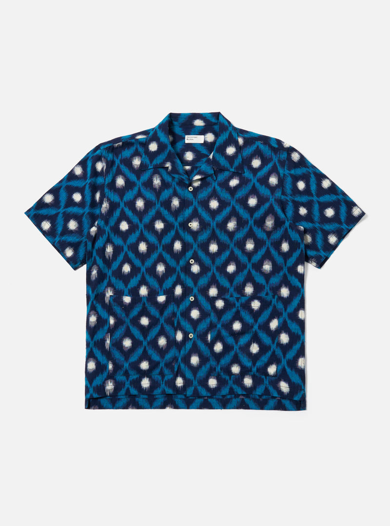 The Indigo Diamond Ikat Deck Shirt is presented as a flat lay against a white background. The shirt features a one-piece collar, short loose sleeves, and two large patch pockets at the lower front. The fabric is decorated with a three-color double ikat diamond pattern in indigo tones.