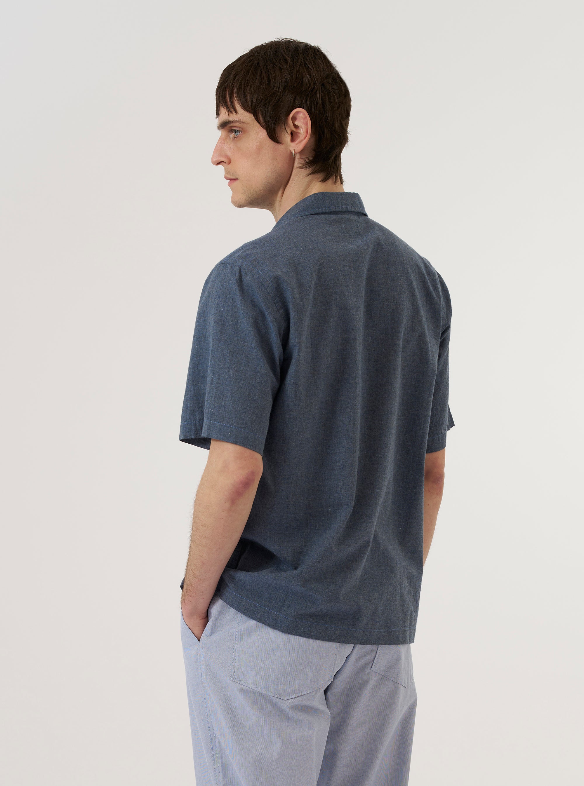A model wears the Indigo Fine Organic Cotton Sal Road Shirt, shown from a rear three-quarter angle against a plain white background. The shirt features a regular fit with short sleeves, a straight hem, and a one-piece collar. The indigo blue organic cotton fabric has a visible marled texture and a lightweight, loose weave.