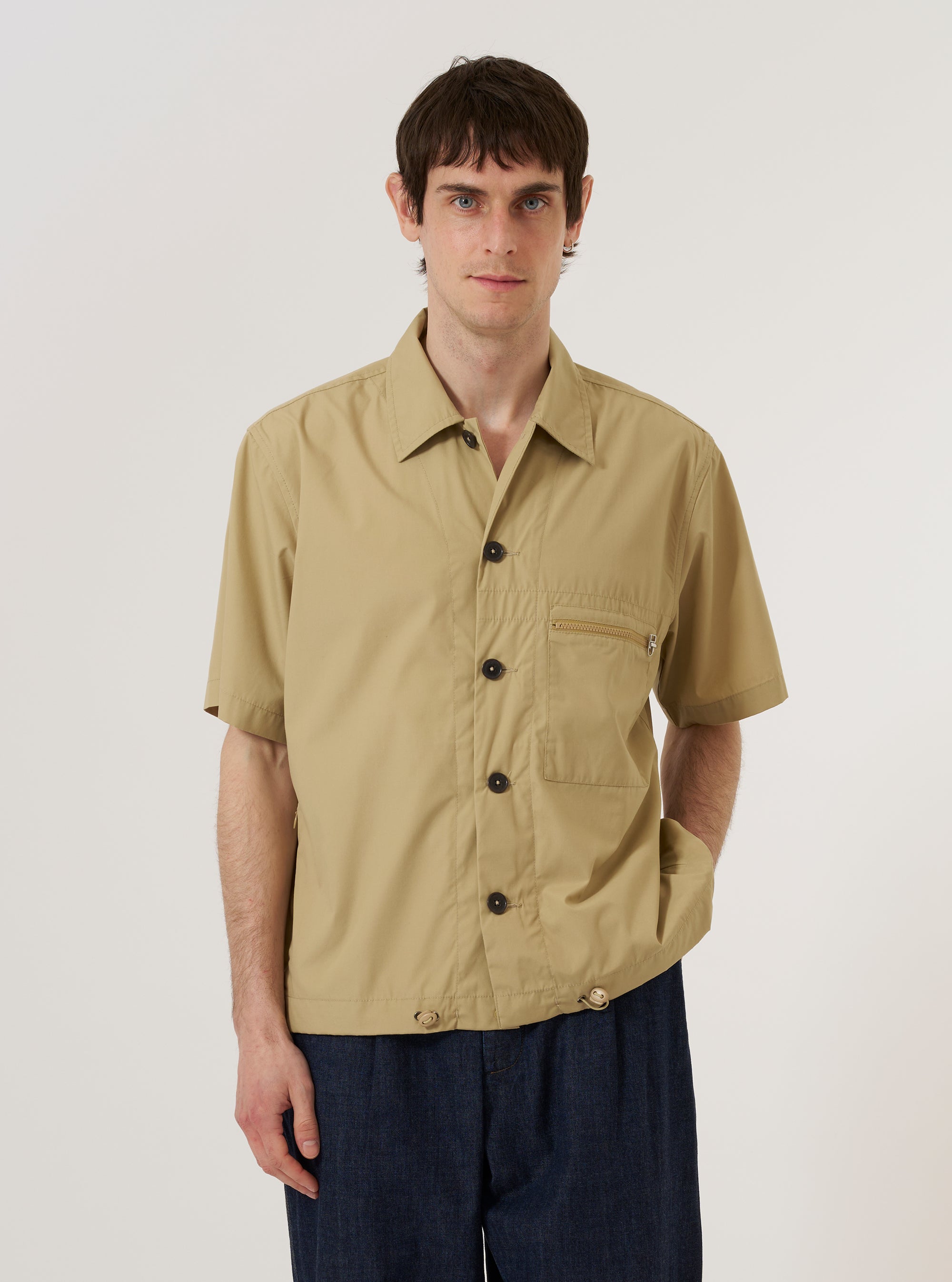 A model wears the Sand Recycled Polytech Z Tech Overshirt over a light-colored top, standing against a plain, light grey background. The utility-style overshirt features a classic collar, a central button-down front, and two large patch pockets at the waist. The sand-colored fabric has a smooth, technical finish.