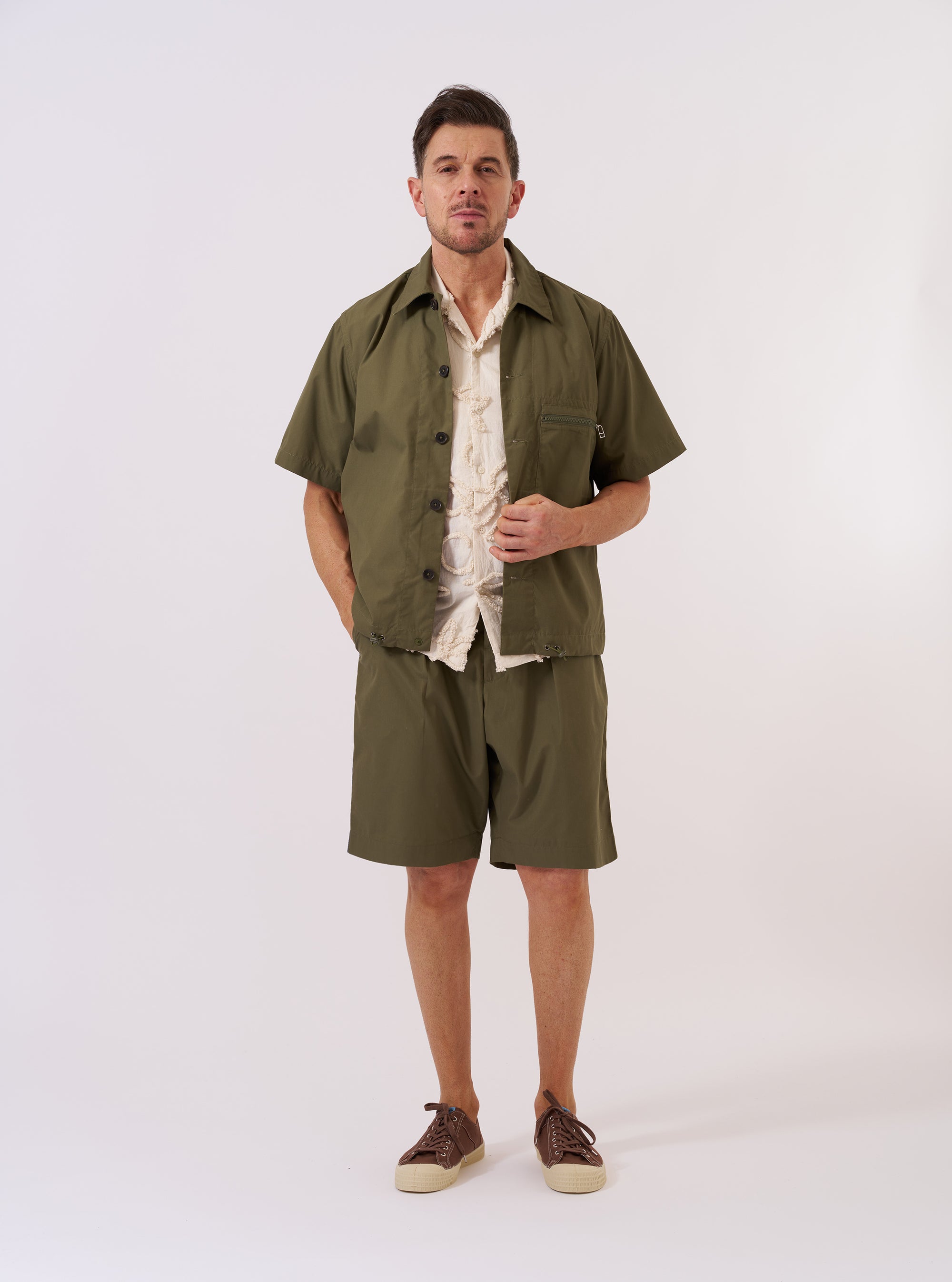 The Olive Recycled Polytech Z Tech Overshirt is shown as a flat lay against a plain white background. The overshirt features a one-piece collar, a central button-down front, and a single reinforced patch pocket on the chest with a vertical zip closure. The olive green fabric has a smooth, technical finish, and the garment is designed with a straight hem and adjustable elasticated drawcords.