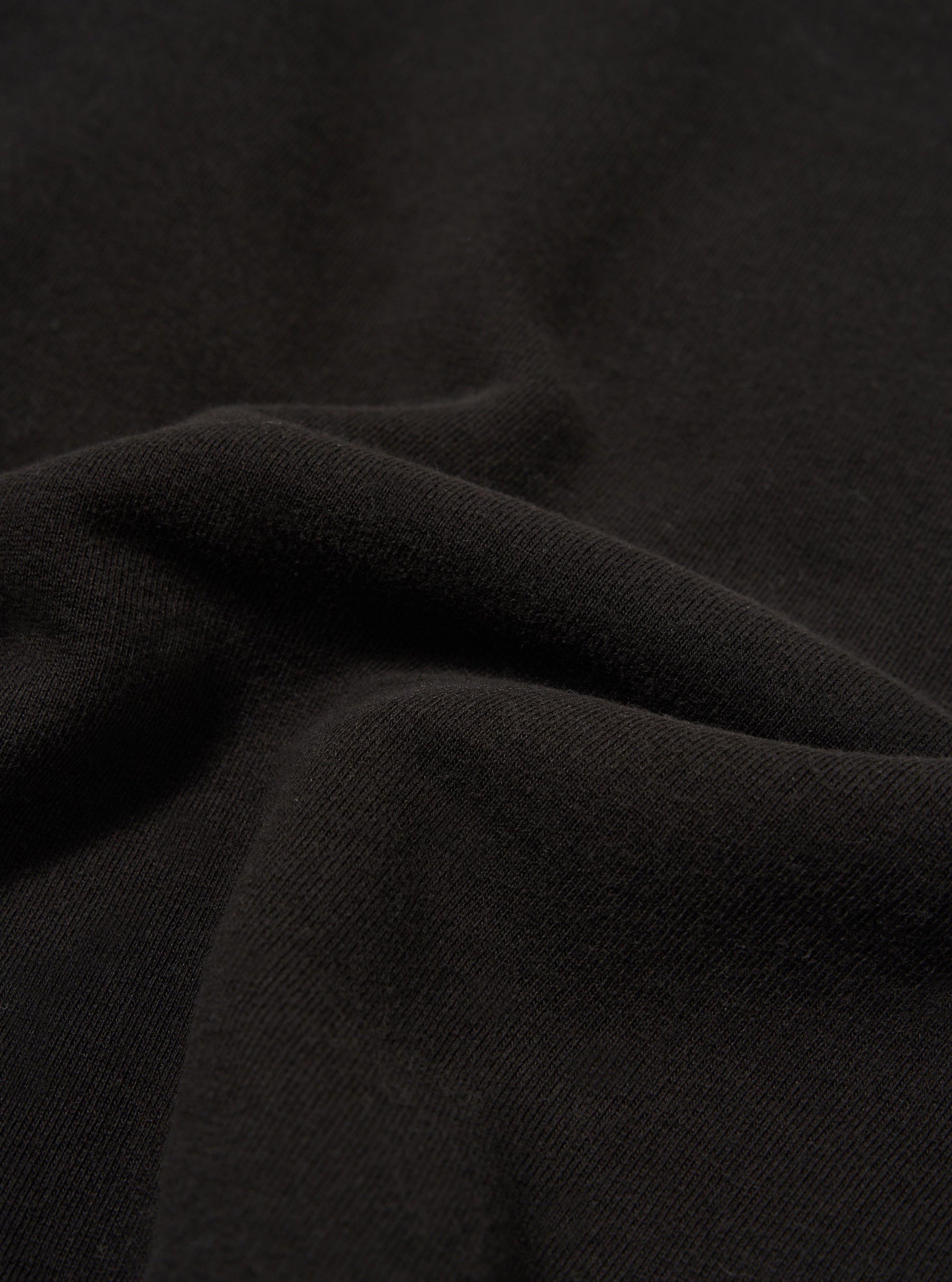 A close-up fabric swatch of the Black Brush Back Sweat Billy Sweat highlights the soft, matte texture of the black cotton material. The image focuses entirely on the fabric's surface, providing a detailed view of its color and composition.