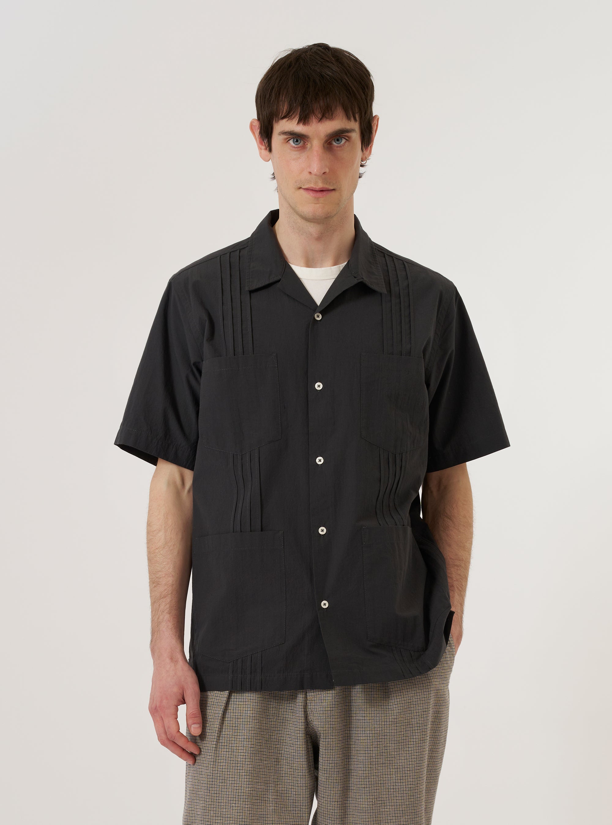 A model wears the Black Tech Cotton S/S Dwight Overshirt against a plain white background. The short-sleeved overshirt features a camp collar, a button-up front, and two large utility-style patch pockets on the chest. The model wears the garment unbuttoned over a white t-shirt, showcasing its relaxed fit and the smooth, technical texture of the black cotton fabric.