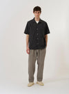 The Black Tech Cotton S/S Dwight Overshirt is shown as a flat lay against a plain white background. The short-sleeved shirt features a camp collar, a central button closure, and four front patch pockets. Two rows of vertical pleats are sewn down the front on either side of the buttons. The fabric is a smooth black tech cotton.