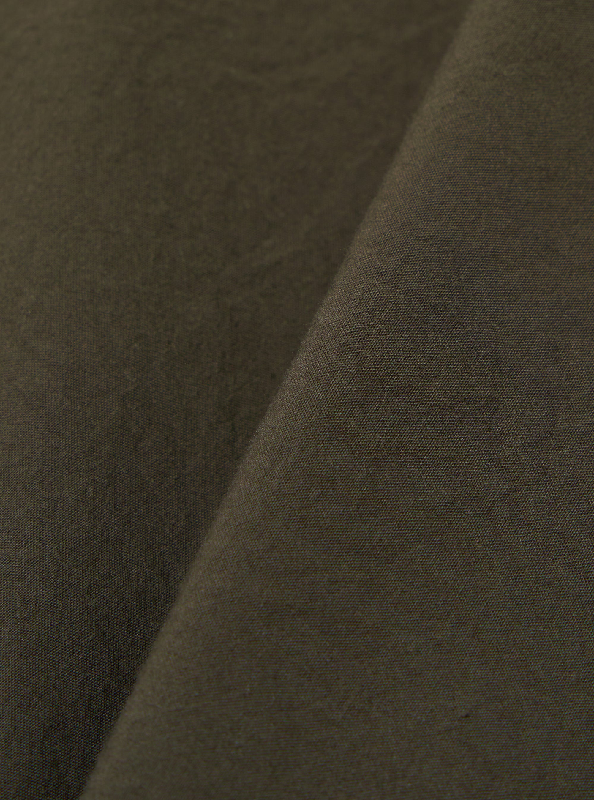 A close-up fabric swatch of the Olive Tech Cotton S/S Dwight Overshirt highlights the olive green color and the smooth texture of the tech cotton fabric. The material fills the frame, providing a detailed view of the garment's construction.