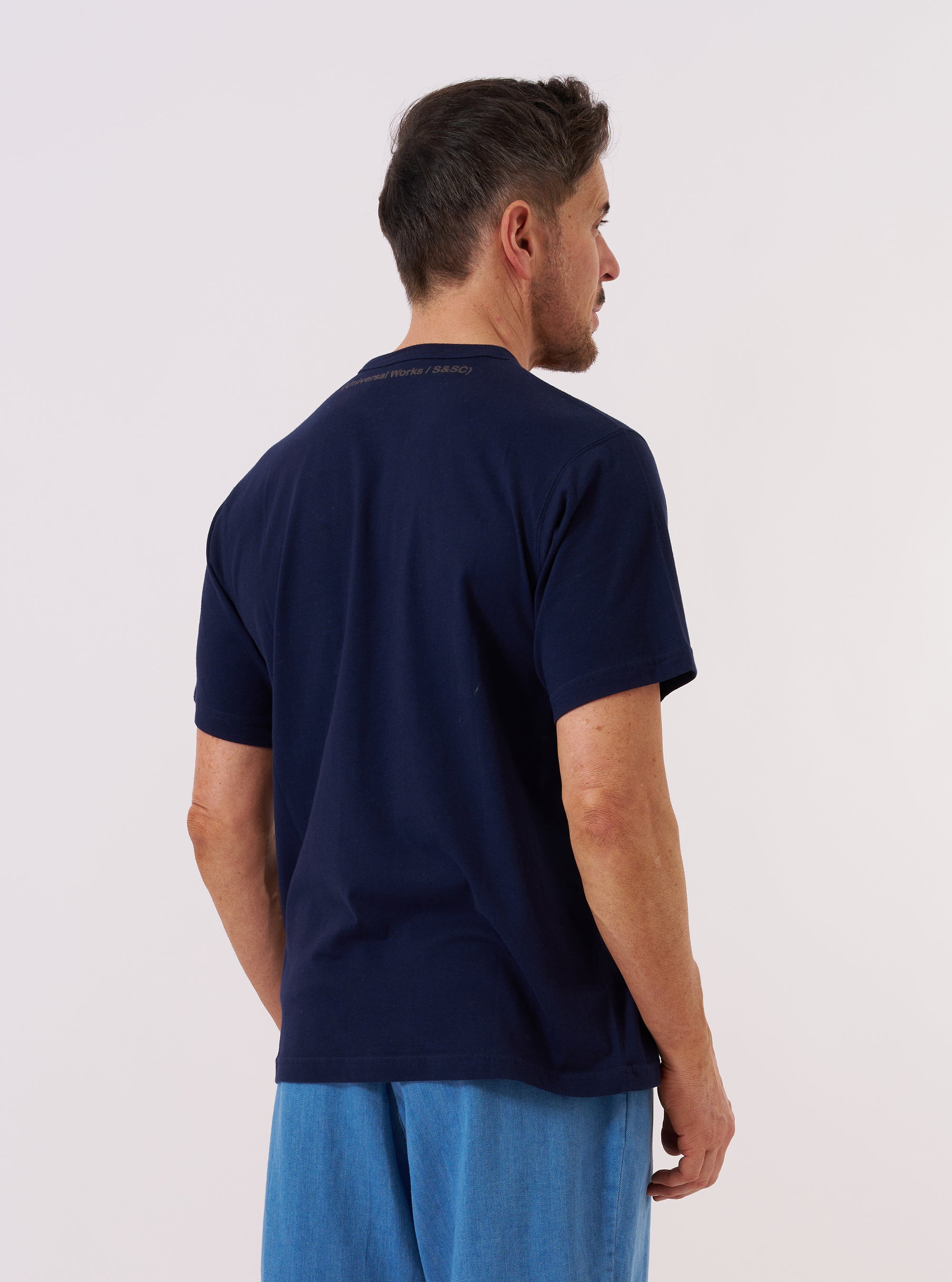 A model wears the Navy Single Jersey 34 Printed Core Tee, shown from a rear angle against a plain white background. The t-shirt features short sleeves and a classic crew neckline, crafted from a navy blue single jersey cotton. The image focuses on the relaxed fit and the clean finish of the garment's back and sleeves.