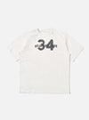 The Ecru Single Jersey 34 Printed Core Tee is shown as a flat lay against a white background. The short-sleeved t-shirt features a ribbed crew neck and a graphic print on the chest. The garment is made from a solid ecru-colored cotton jersey.
