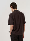 A model wears the Brown Diamond Dobby Deck Shirt over a light-colored top, shown from a rear three-quarter angle against a plain white background. The shirt features a relaxed fit, a straight hem, and a distinctive diamond-shaped dobby weave throughout the brown cotton fabric. The back of the garment shows a clean construction with a simple yoke and a single vertical seam.
