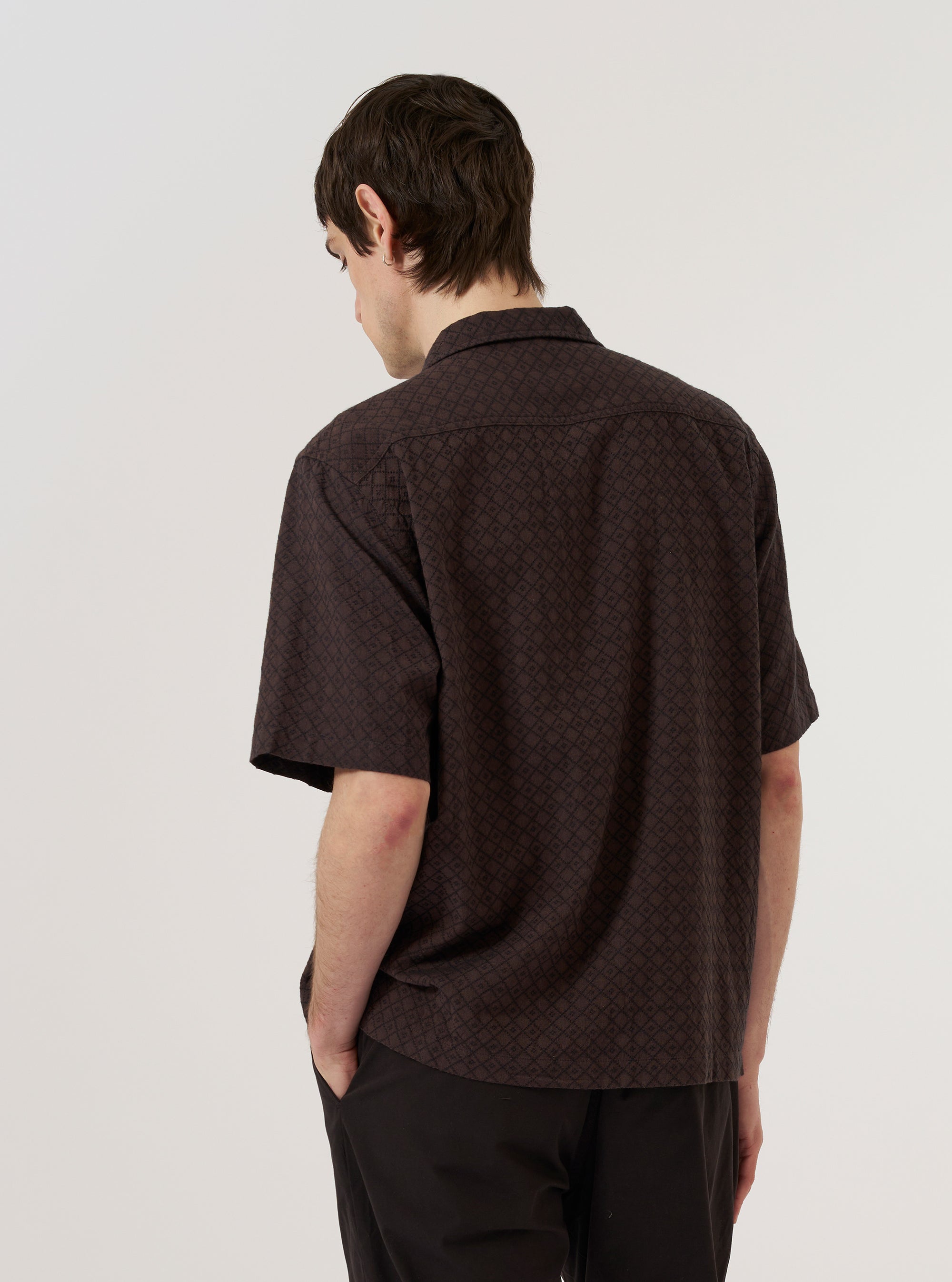 A model wears the Brown Diamond Dobby Deck Shirt over a light-colored top, shown from a rear three-quarter angle against a plain white background. The shirt features a relaxed fit, a straight hem, and a distinctive diamond-shaped dobby weave throughout the brown cotton fabric. The back of the garment shows a clean construction with a simple yoke and a single vertical seam.