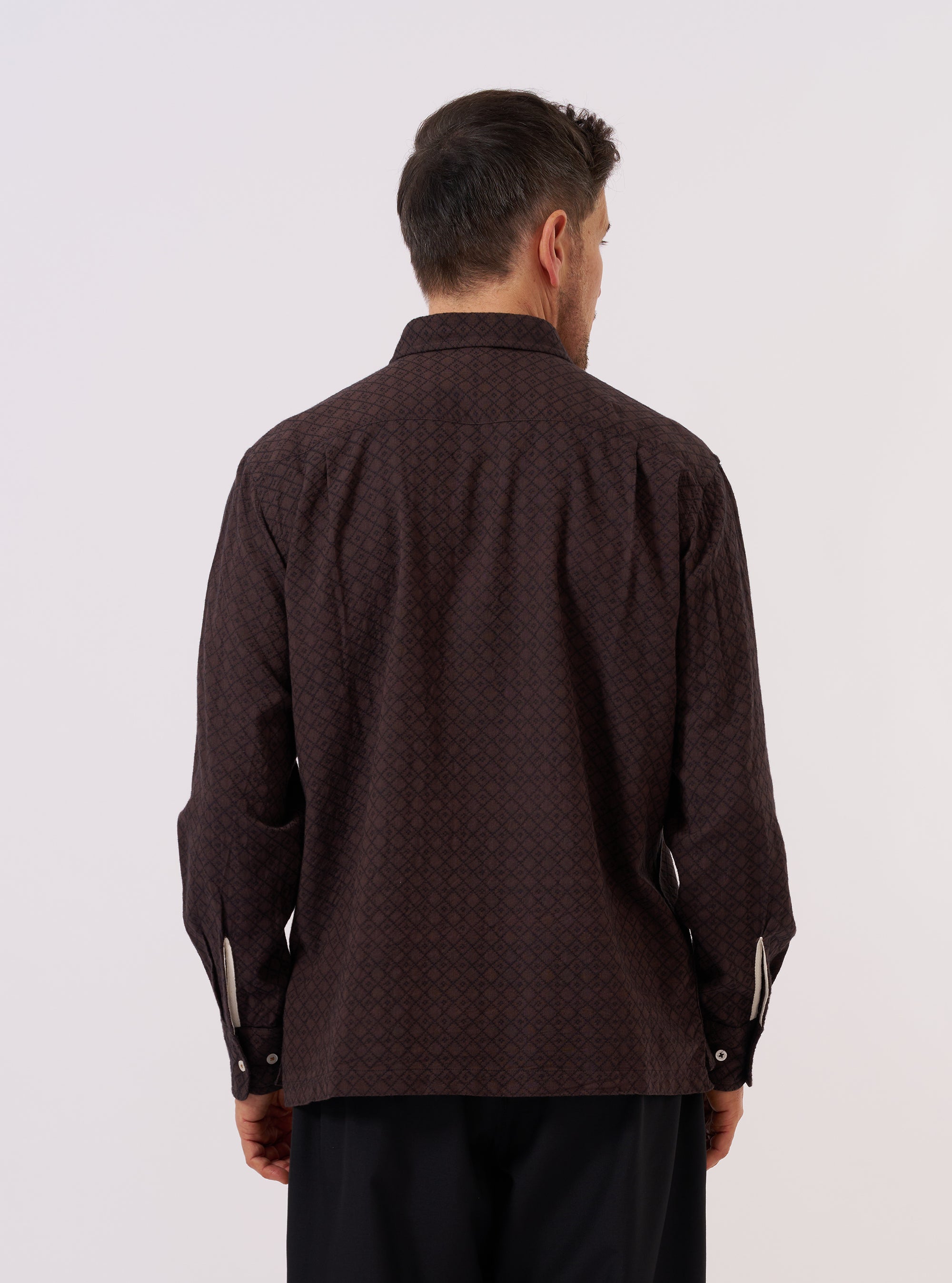 A model wears the Brown Diamond Dobby Lucky Shirt, shown from a rear three-quarter angle against a plain, neutral studio background. The shirt features a relaxed silhouette, buttoned cuffs, and a straight hem. The brown cotton fabric is characterized by a distinct and consistent textured diamond dobby pattern throughout.