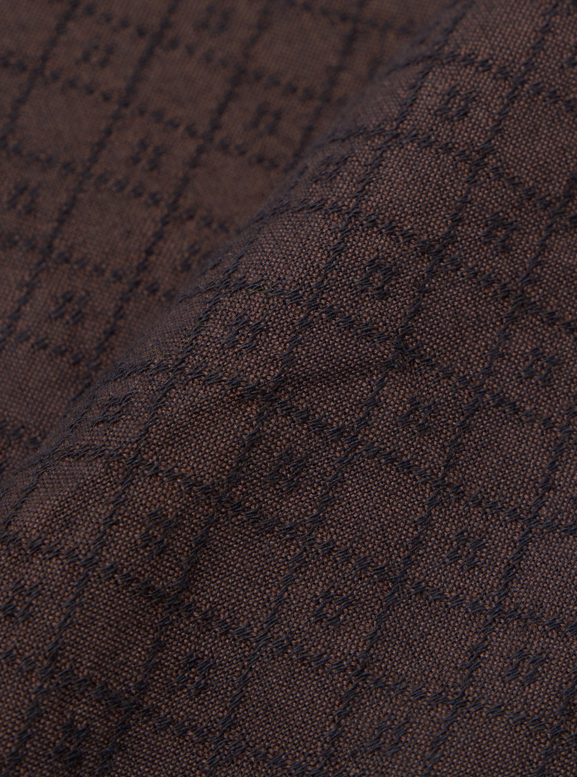A close-up fabric swatch of the Brown Diamond Dobby Lucky Shirt displays a detailed view of the brown cotton material. The image highlights the intricate, geometric diamond dobby weave and the textured surface of the fabric.