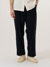 A model wears the Black Organic Fine Cord Oxford Pant, standing against a plain white background. The image shows a front view of the trousers, highlighting the loose, wide-leg fit, the single pleat at the front, and the elasticated drawstring waistband. The black organic fine corduroy fabric has a visible vertical ribbed texture. The model is wearing a light-colored top tucked in to reveal the waistband and pleat details.